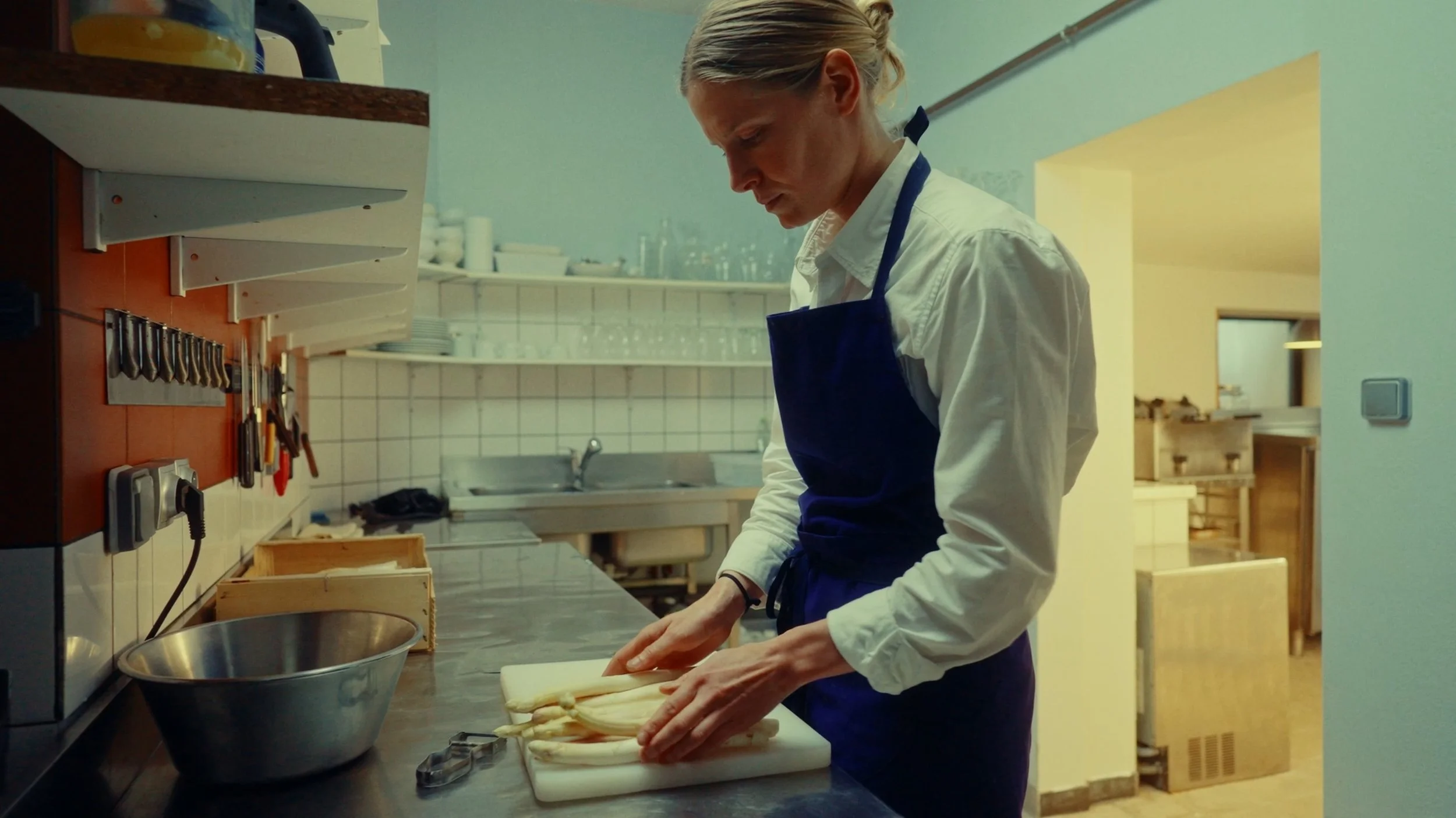 Une femme en uniforme de cuisine, portant un tablier bleu foncé, coupe du pain ou de la pâte dans une cuisine professionnelle.