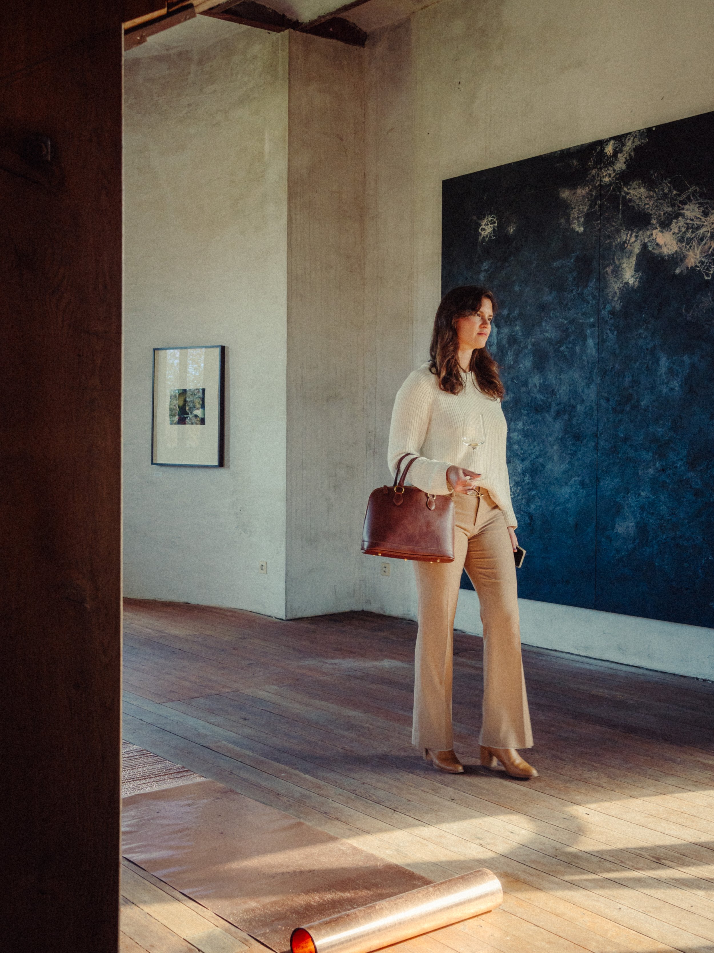 Femme debout dans une galerie d'art, portant un pull blanc, un pantalon beige, des bottes marron, et tenant un verre de vin blanc, avec un sac à main marron.