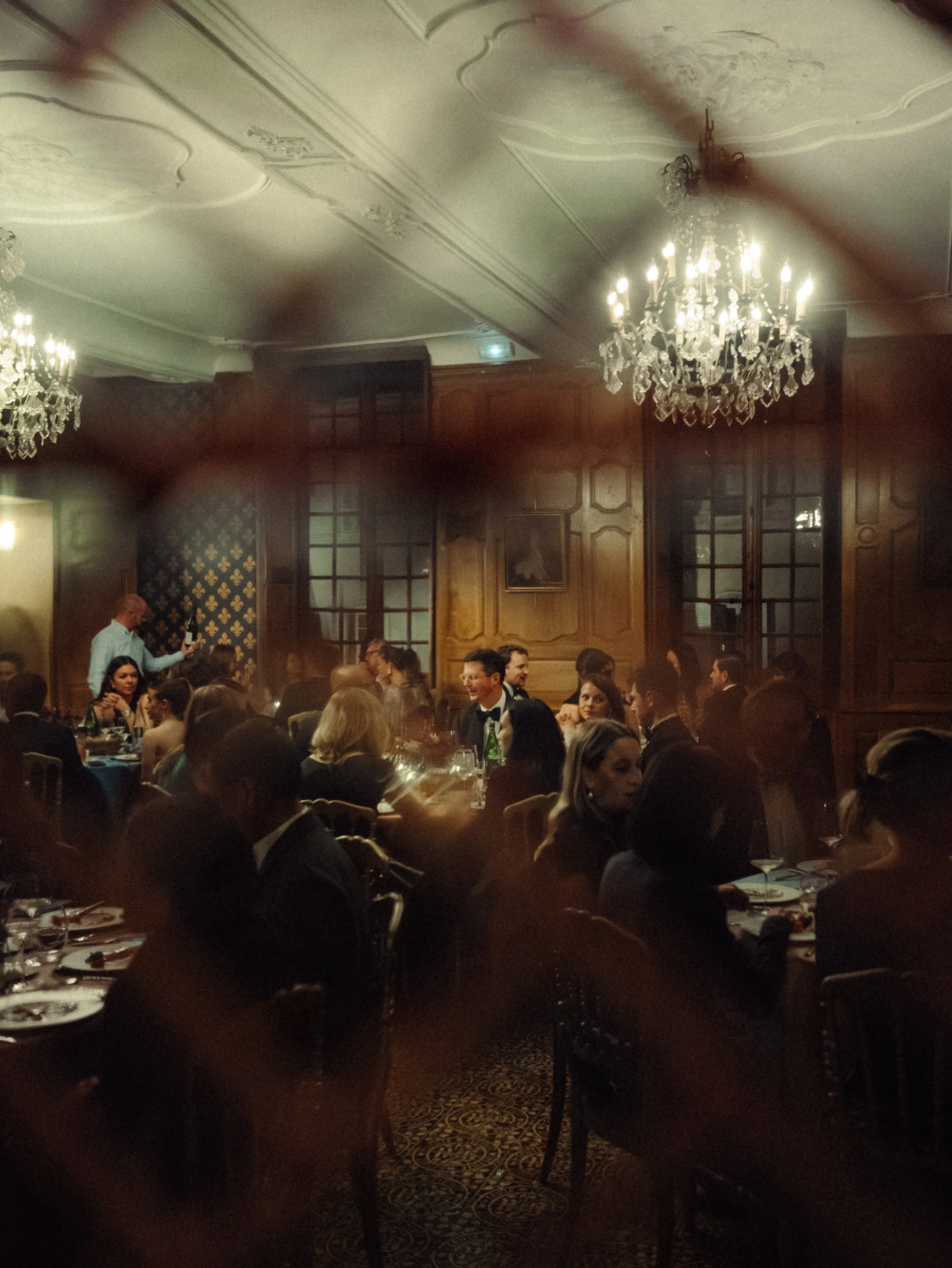 Un dîner élégant dans une salle somptueuse avec des lustres en cristal, des invités en tenue formelle assis à des tables, probablement lors d'un événement ou d'une réception.