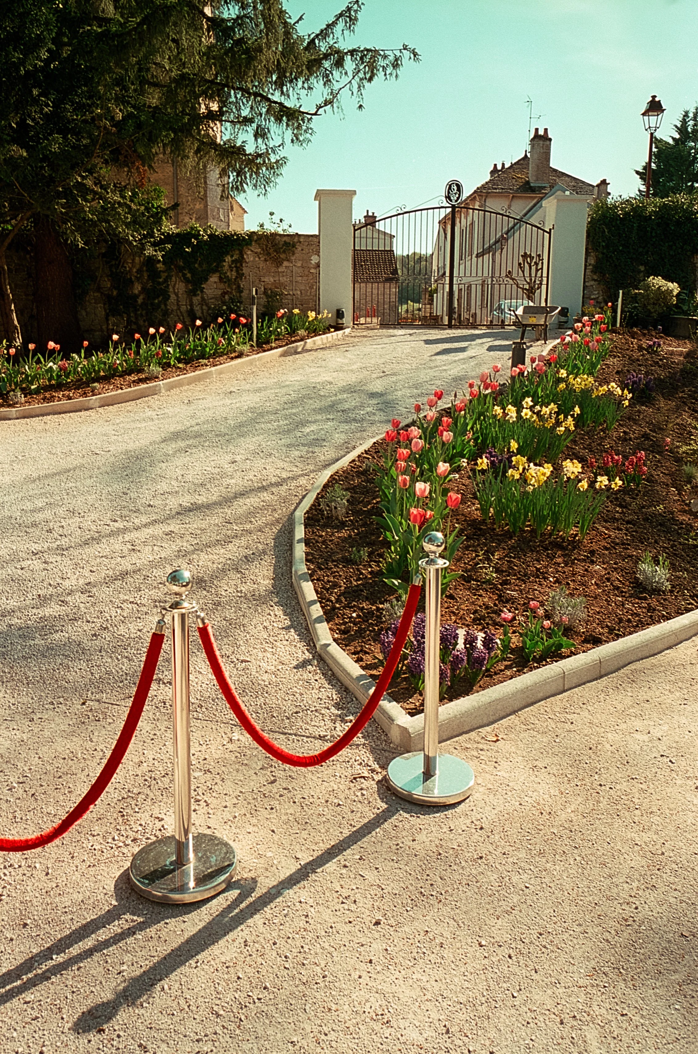 Jardin de fleurs autour d'une entrée avec barrière rouge et poutres en acier inoxydable, voie d'accès pavée, maison en arrière-plan avec une porte en fer forgé.