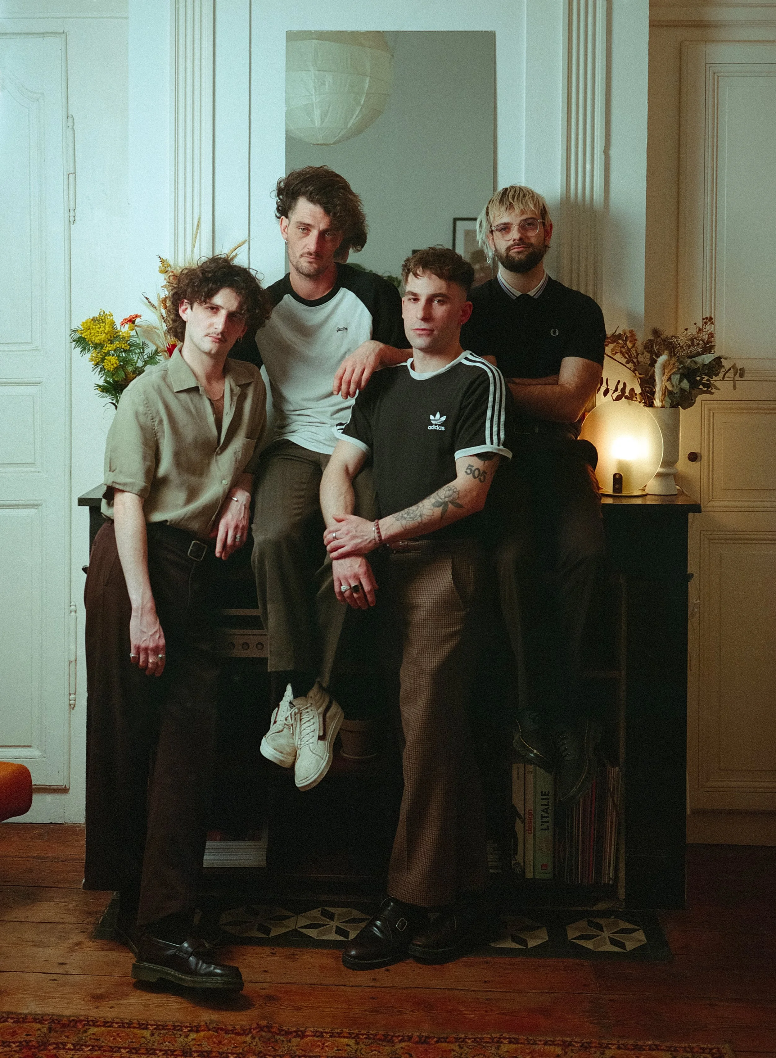 Groupe de cinq jeunes hommes posant dans un intérieur, certains assis, d'autres debout, avec des fleurs et une lampe derrière eux.