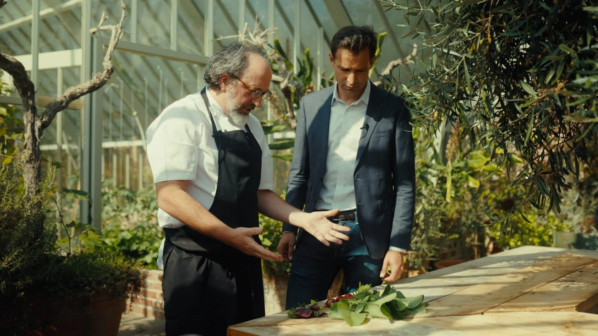 Deux hommes discutent dans une serre ou un espace vert, en regardant un tableau de composition florale ou de plantes sur une table en bois.