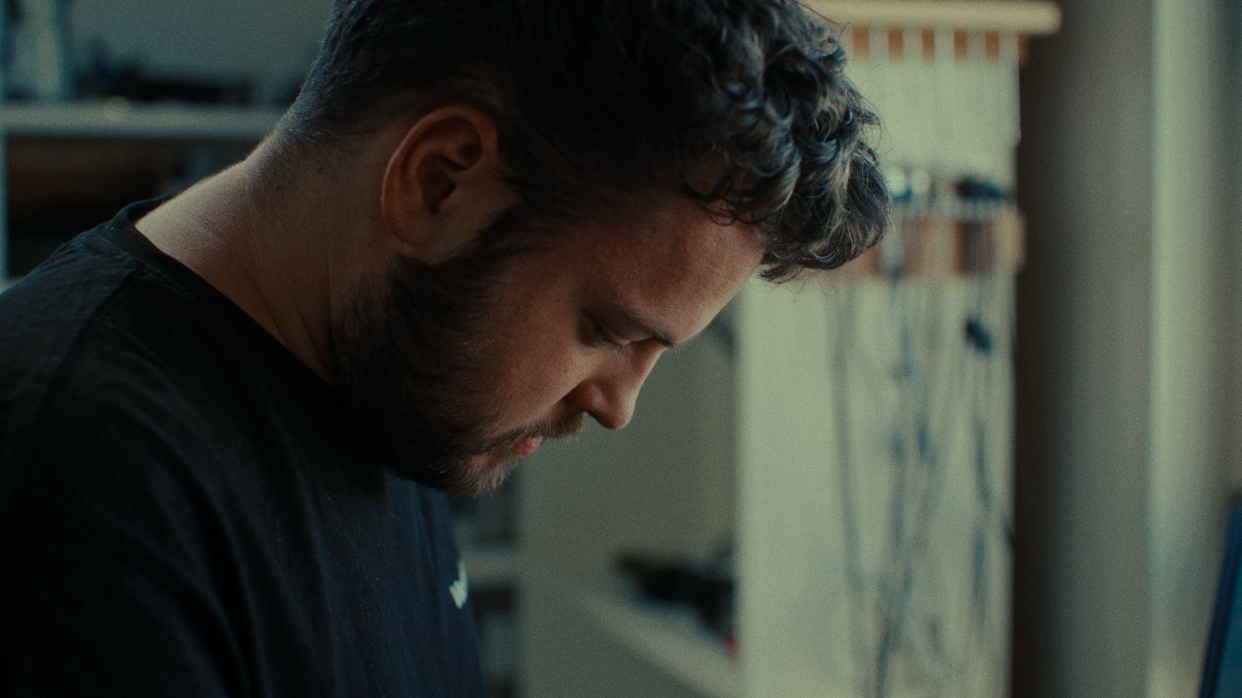 Un homme avec une barbe et des cheveux bouclés regarde vers le bas dans une pièce avec des étagères et des objets en arrière-plan.