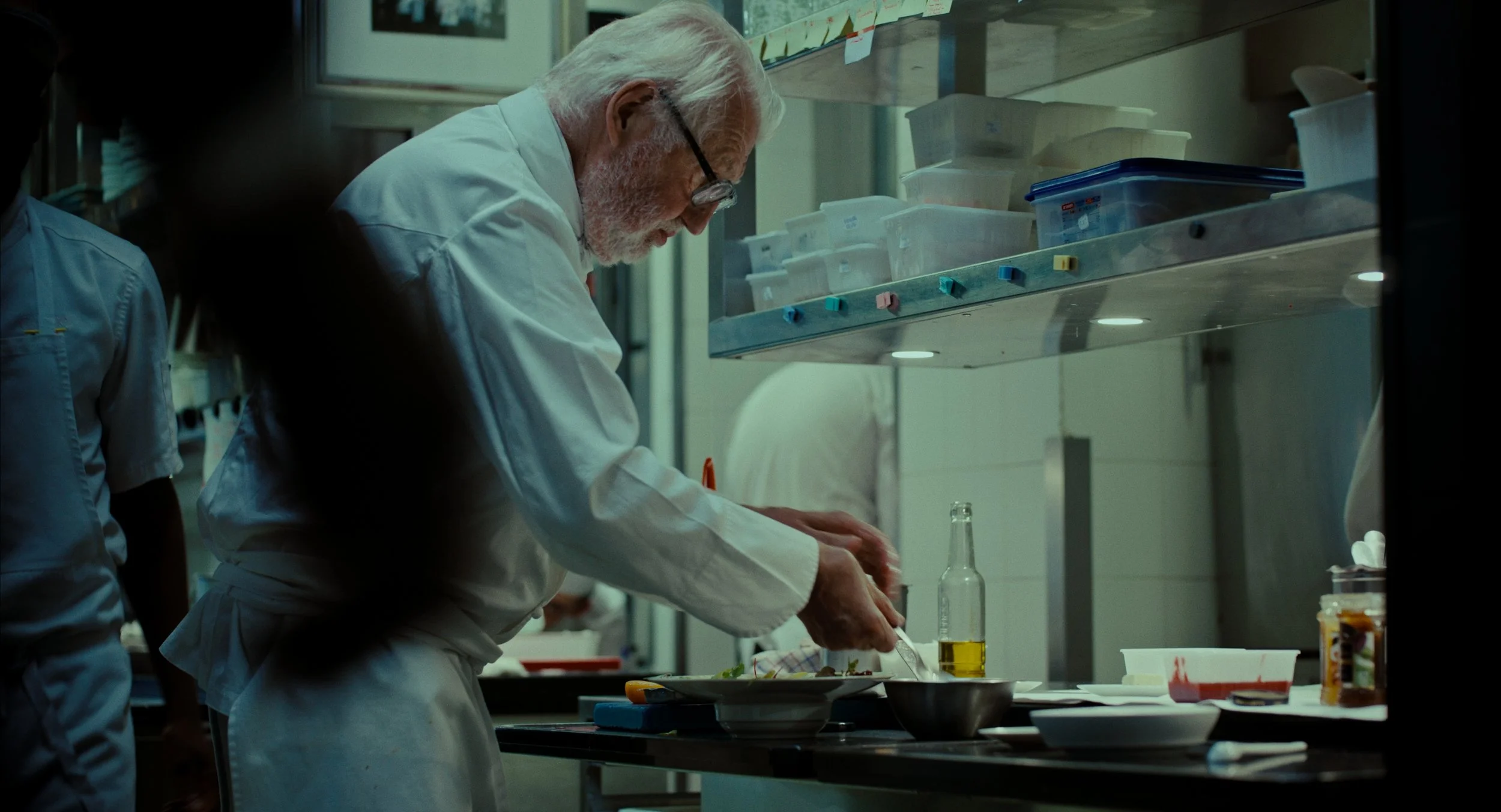 Un chef cuisinier âgé en blanc préparant un plat dans une cuisine professionnelle, avec des contenants transparents et des fournitures culinaires sur une étagère en arrière-plan.
