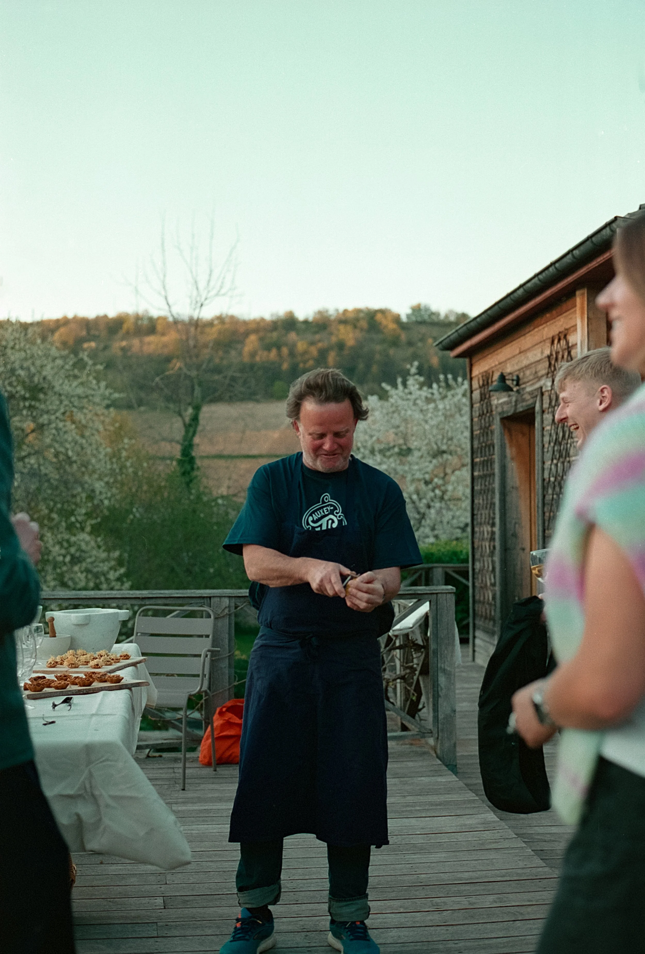 Un chef en costume noir et avec une barbe, souriant et en train de préparer quelque chose lors d'une réunion en plein air, avec des personnes autour, un ciel clair en arrière-plan et une structure en bois à côté.