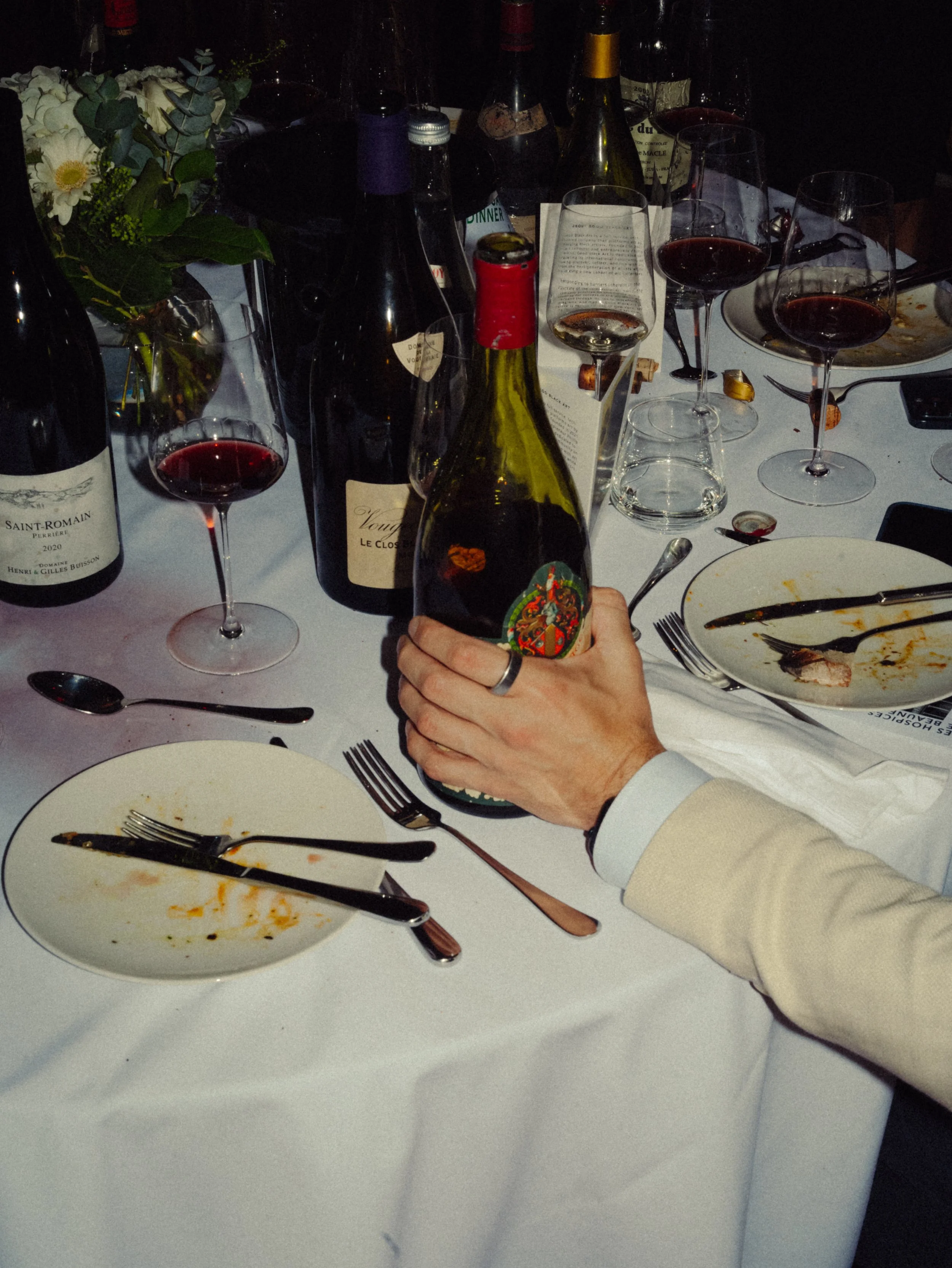 Table avec des verres de vin rouge, des bouteilles de vin, des assiettes vides, des couverts, et une main tenant une bouteille de vin sur fond de soirée ou dîner.