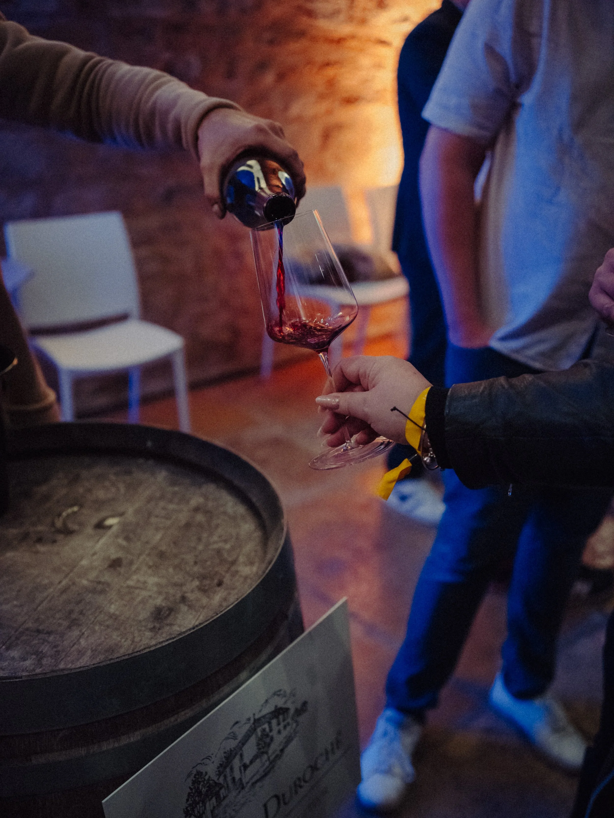 Une personne verse du vin rouge dans un verre lors d'une dégustation dans une cave en pierre, avec d'autres personnes autour.