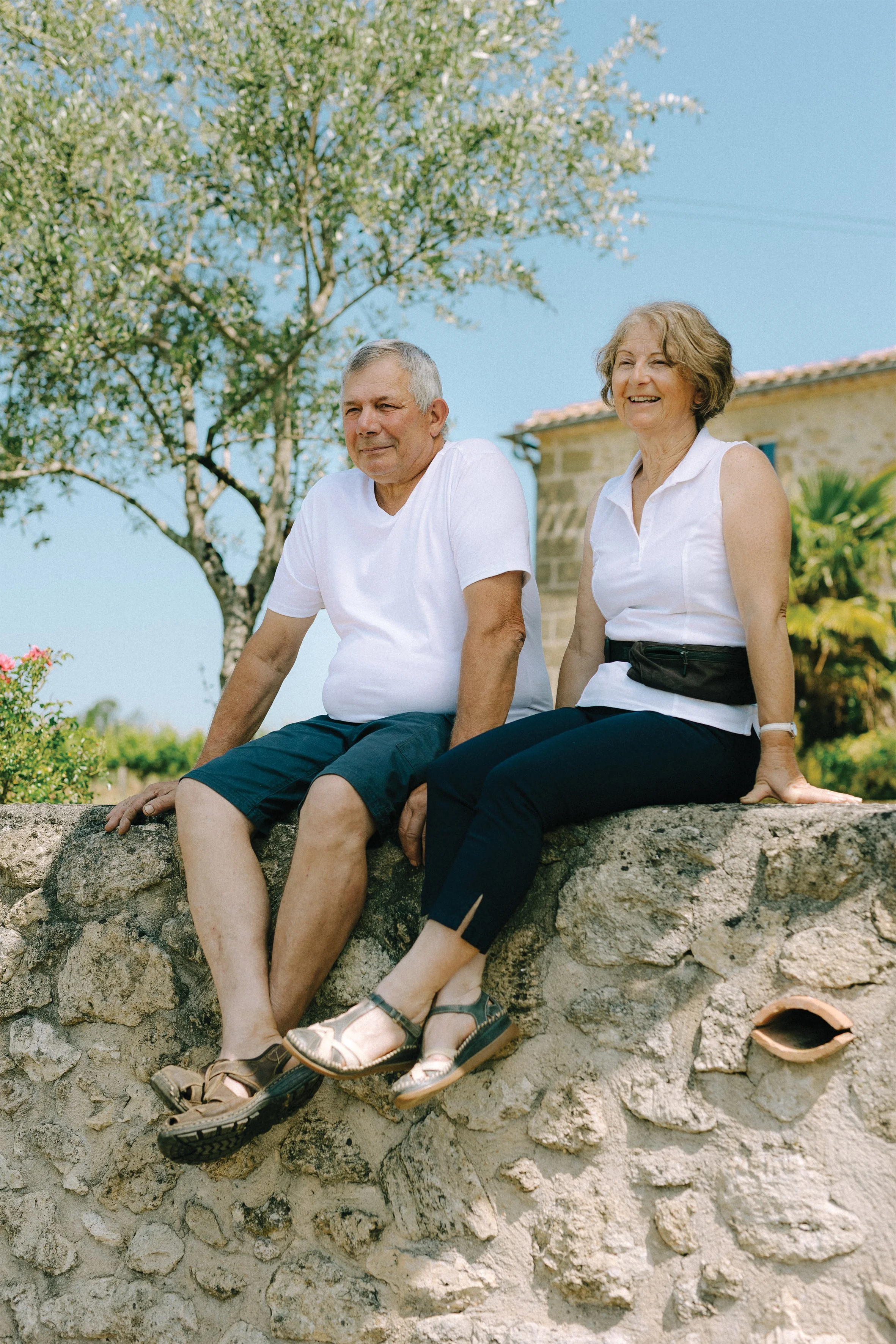 Deux personnes âgées, un homme et une femme, assises sur un mur en pierre sous un arbre, souriantes, par une journée ensoleillée.