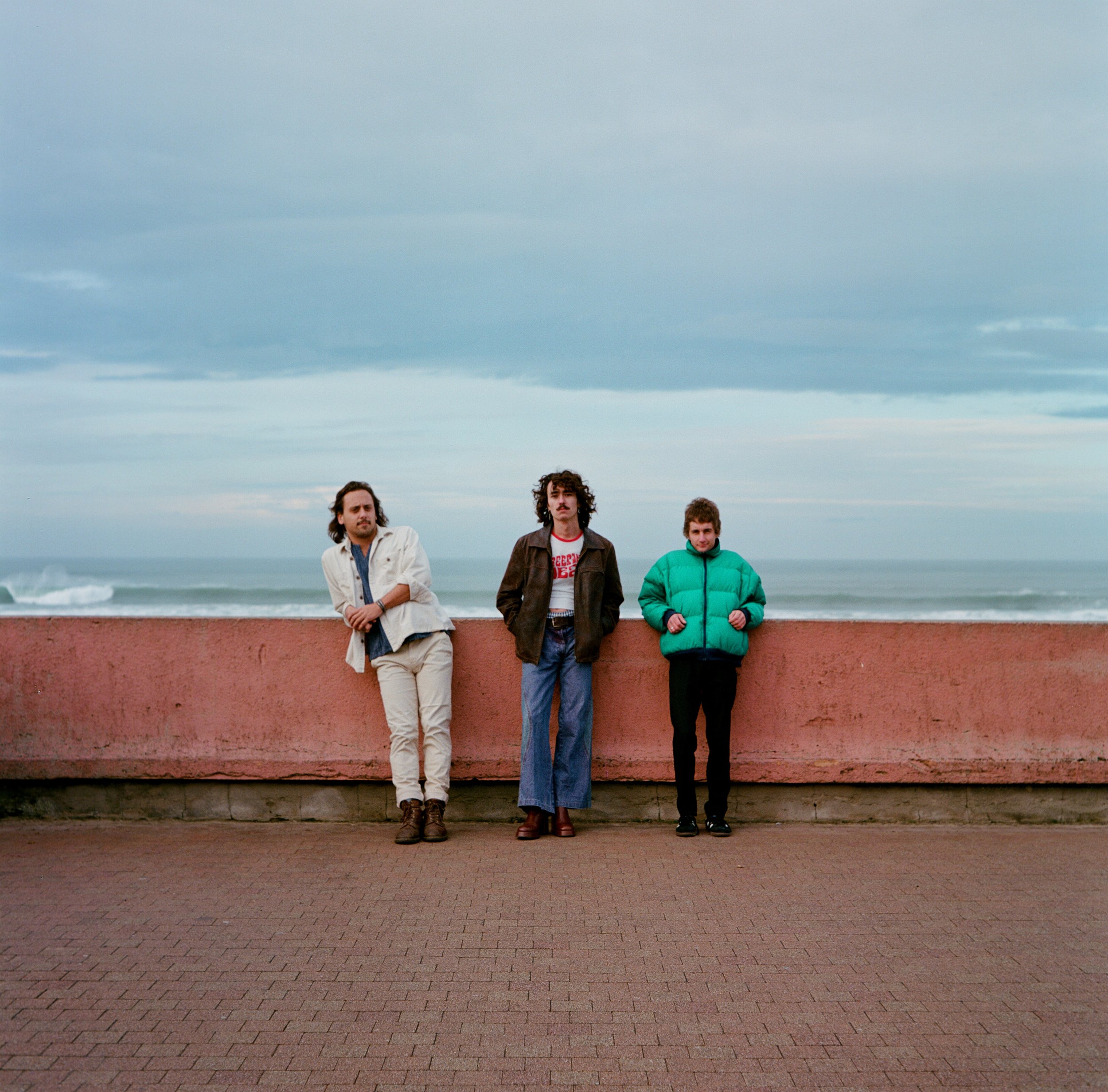 Trois jeunes hommes debout devant une barrière en bord de mer, vêtus de vêtements décontractés, avec un ciel nuageux en arrière-plan.