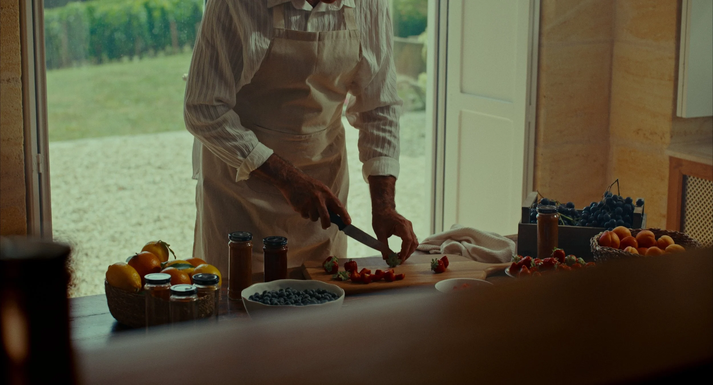 Une personne prépare des fraises sur une planche à découper, entourée de fruits comme des pêches, des myrtilles, et des abricots dans une cuisine lumineuse.