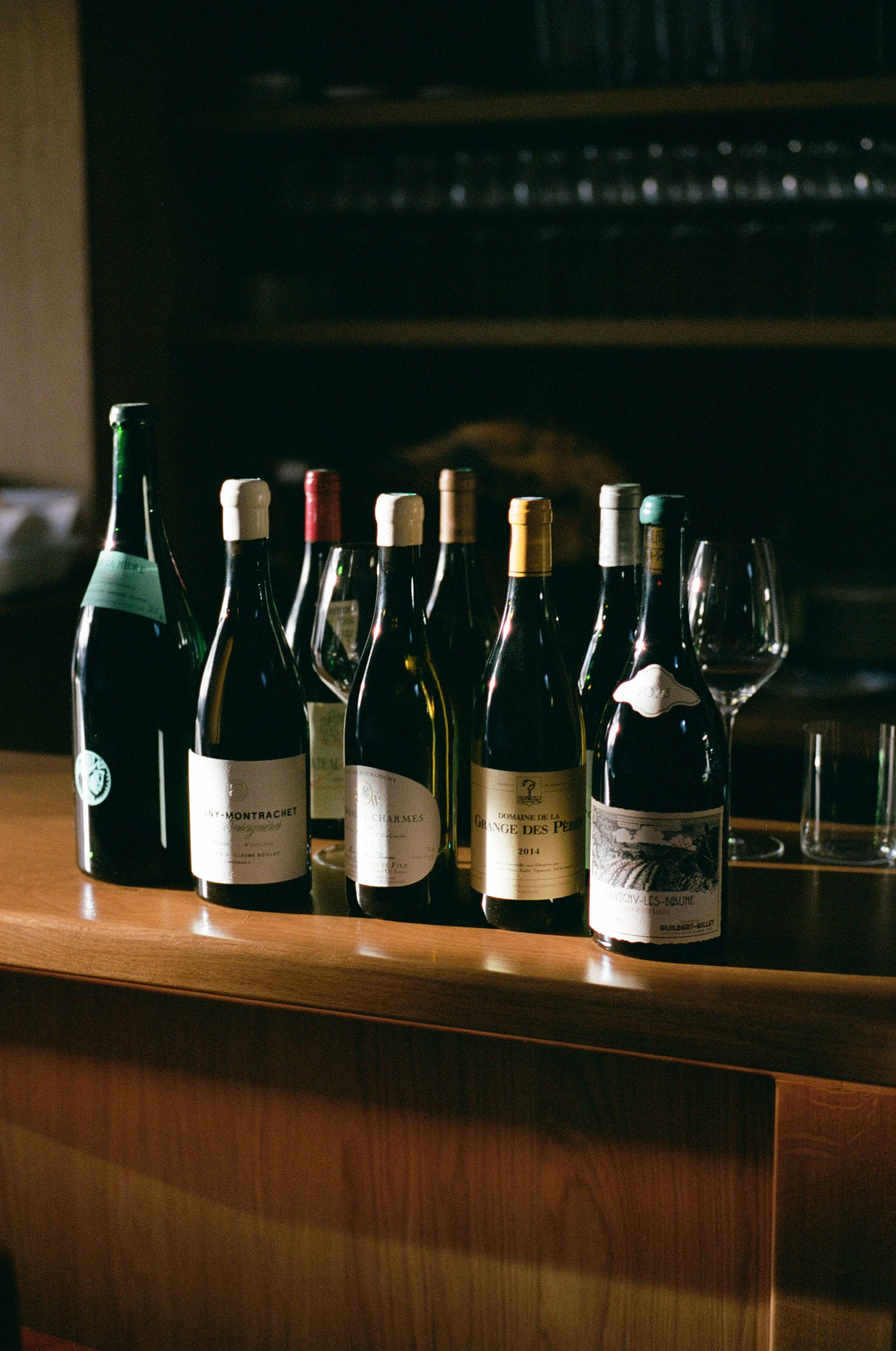 Plusieurs bouteilles de vin alignées sur un comptoir en bois, avec des verres à vin vides en arrière-plan.