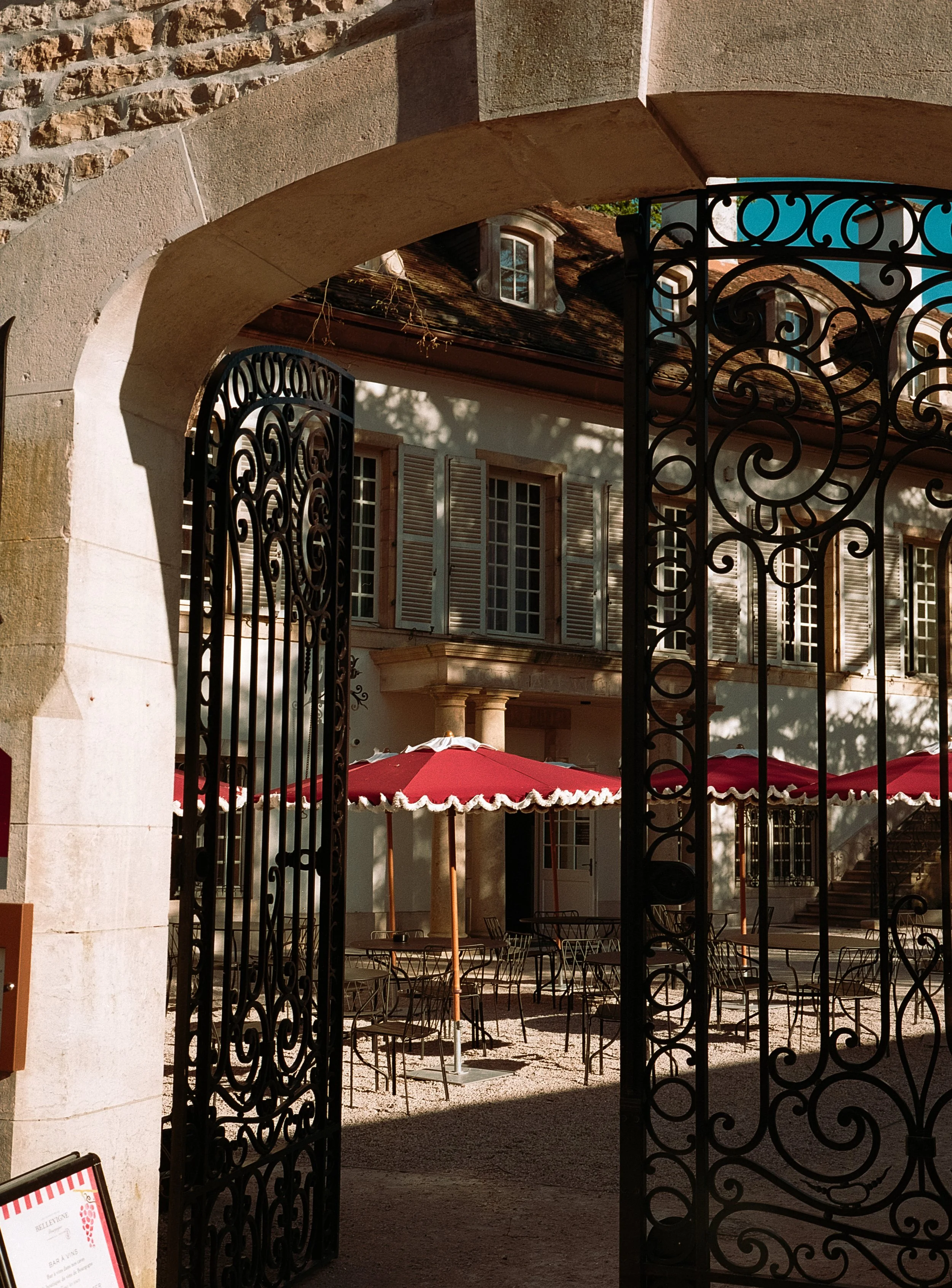 Une cour extérieure avec des parasols rouges, des tables et des chaises en métal, vue à travers une porte en fer forgé, bâtiment en arrière-plan avec fenêtres à volets.