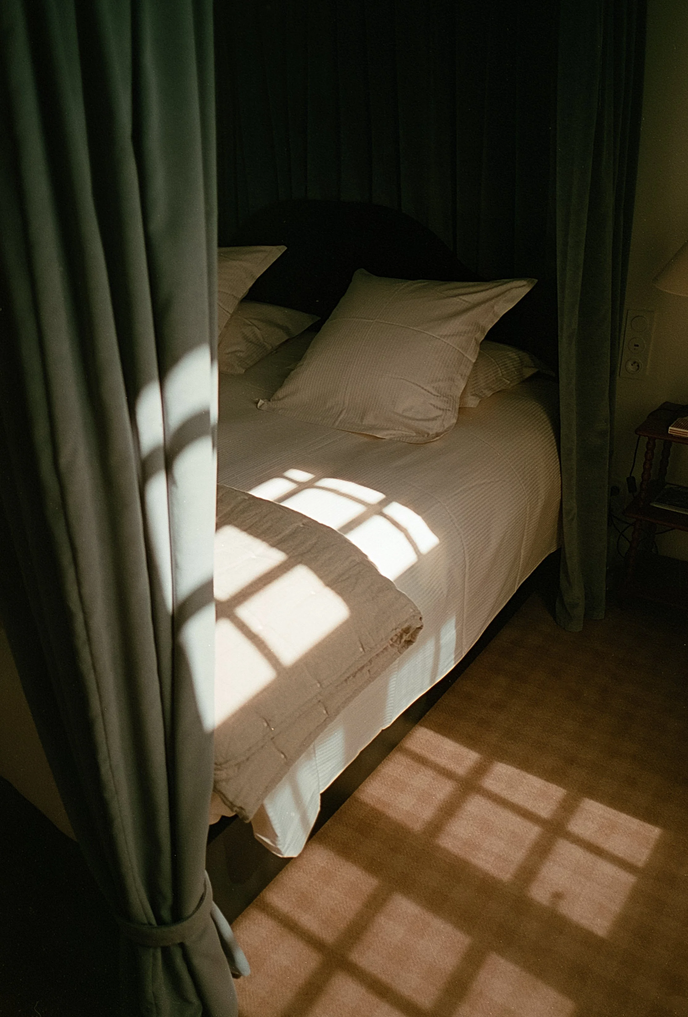 Lit fait de draps et de coussins, avec des rideaux en velours vert foncé et des rayons de soleil créant un motif carré sur le sol en bois.