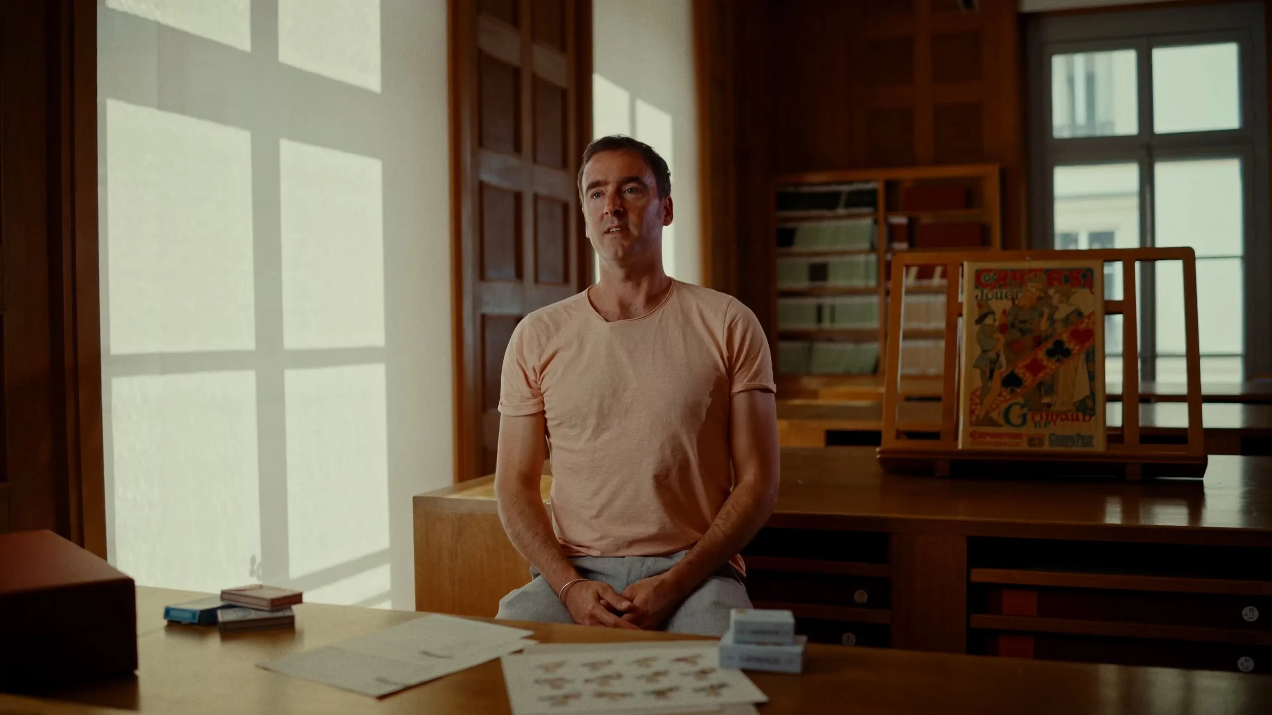 Jeune homme assis dans une pièce avec mobilier en bois, voyant de grande fenêtre, affiches et documents sur une table.