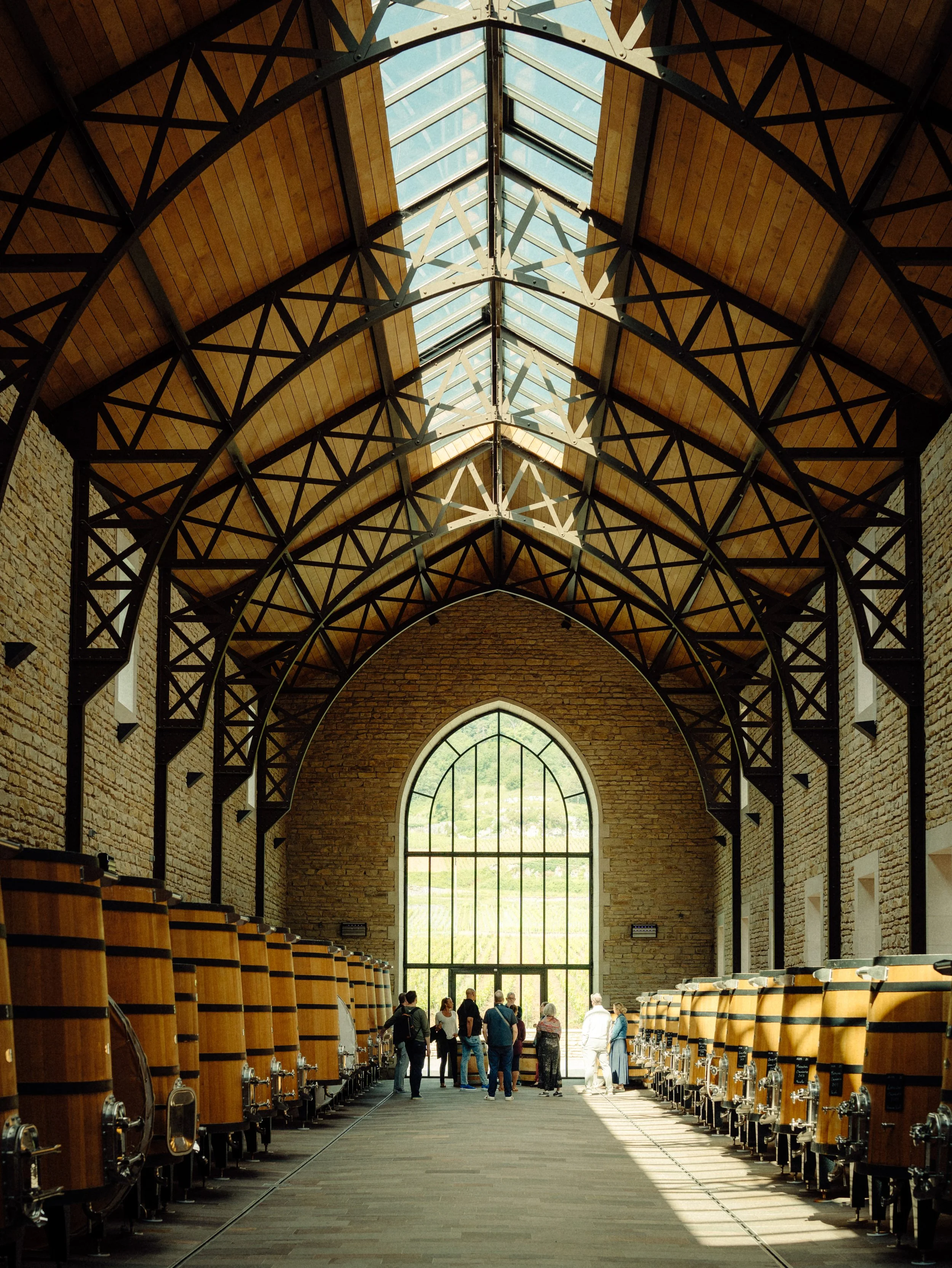 Une salle de dégustation de vin avec des fûts en bois alignés de chaque côté, un grand fenêtre en arc laissant entrer la lumière, et un groupe de personnes au centre discutant.
