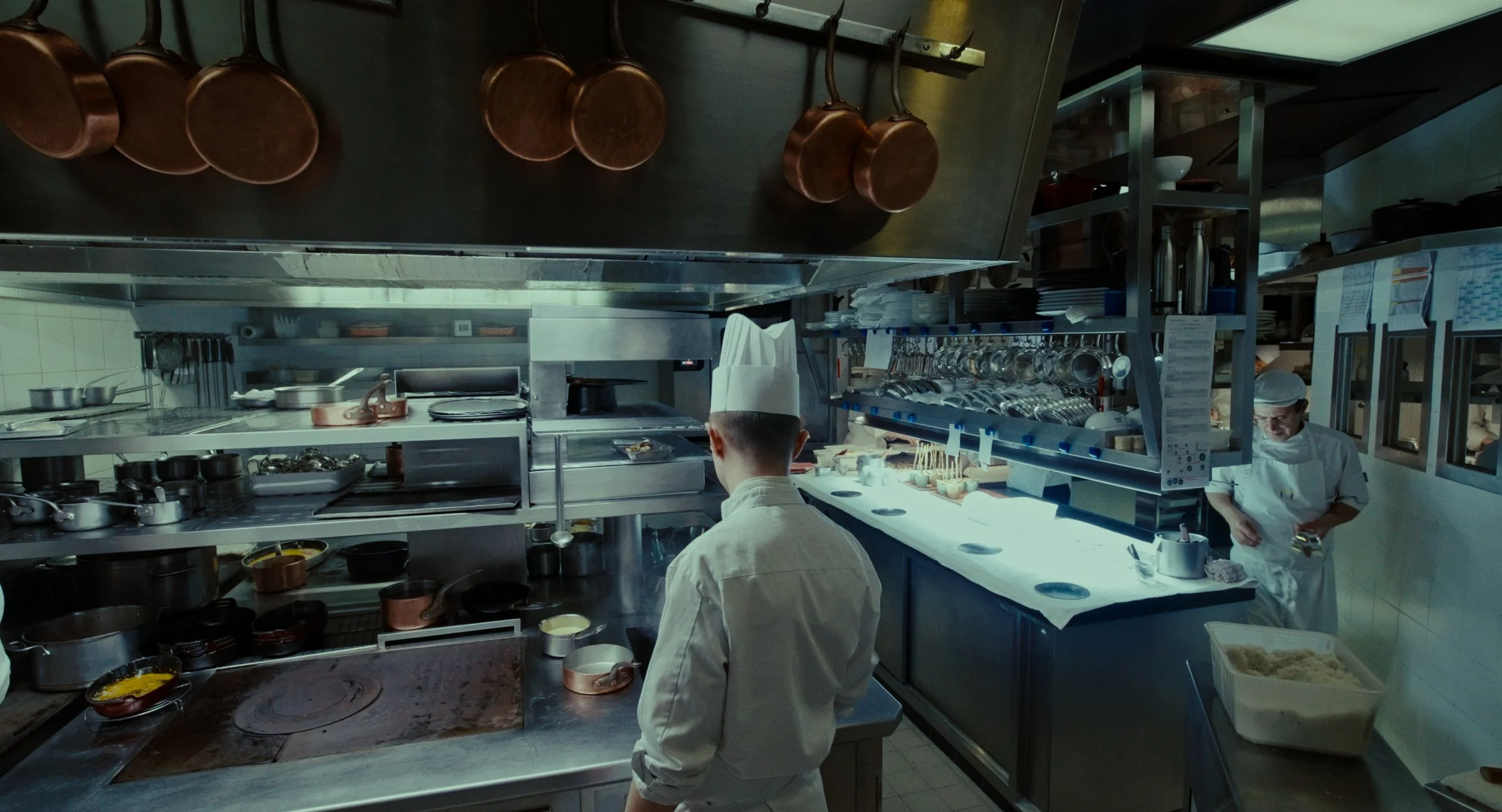 Cuisine professionnelle avec deux chefs en uniforme, étagères de casseroles, planche de travail éclairée, ustensiles et ingrédients