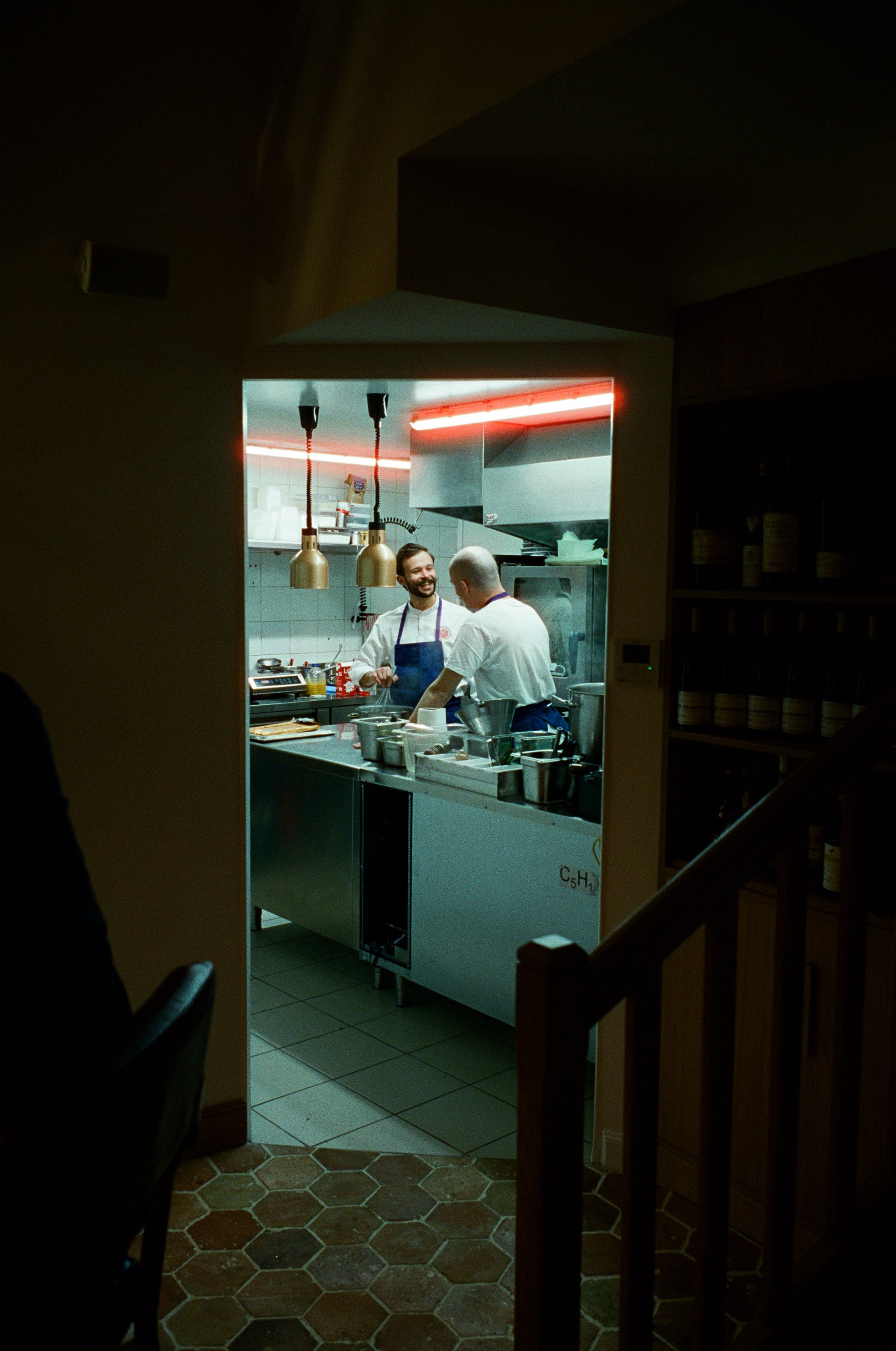 Deux chefs qui discutent dans une cuisine professionnelle, vue à travers une porte ou une ouverture dans un mur.