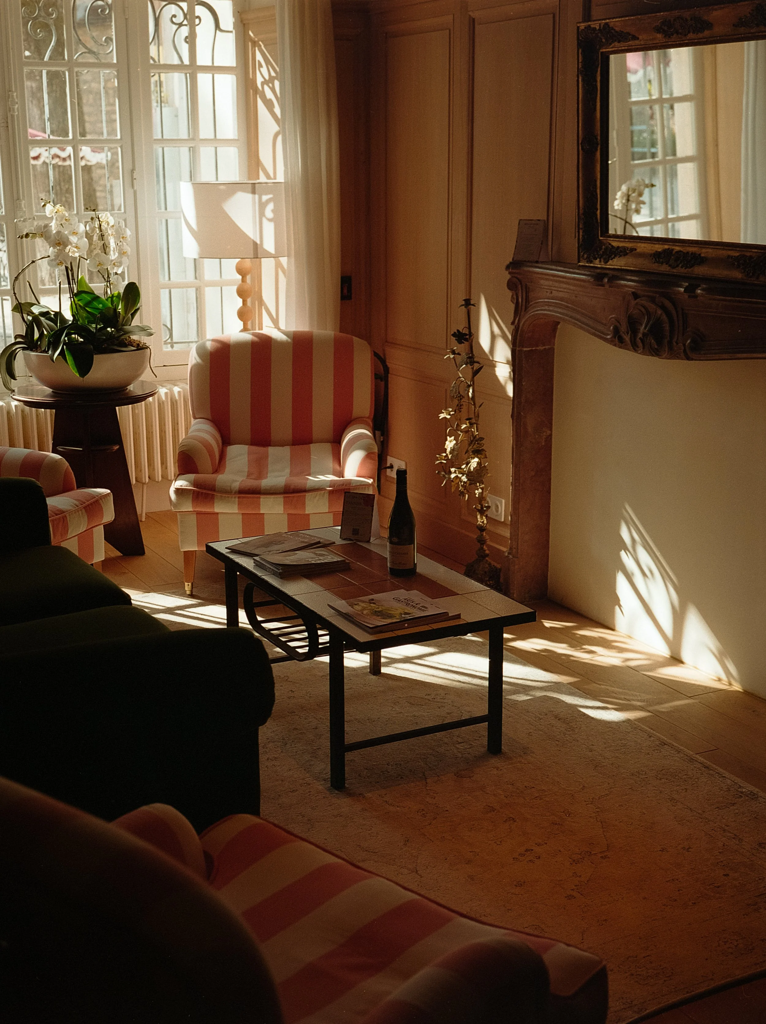Intérieur d'un salon avec fauteuils à rayures, table basse avec des magazines, une bouteille de vin, une cheminée en marbre, un grand miroir, et une plante orchidée.