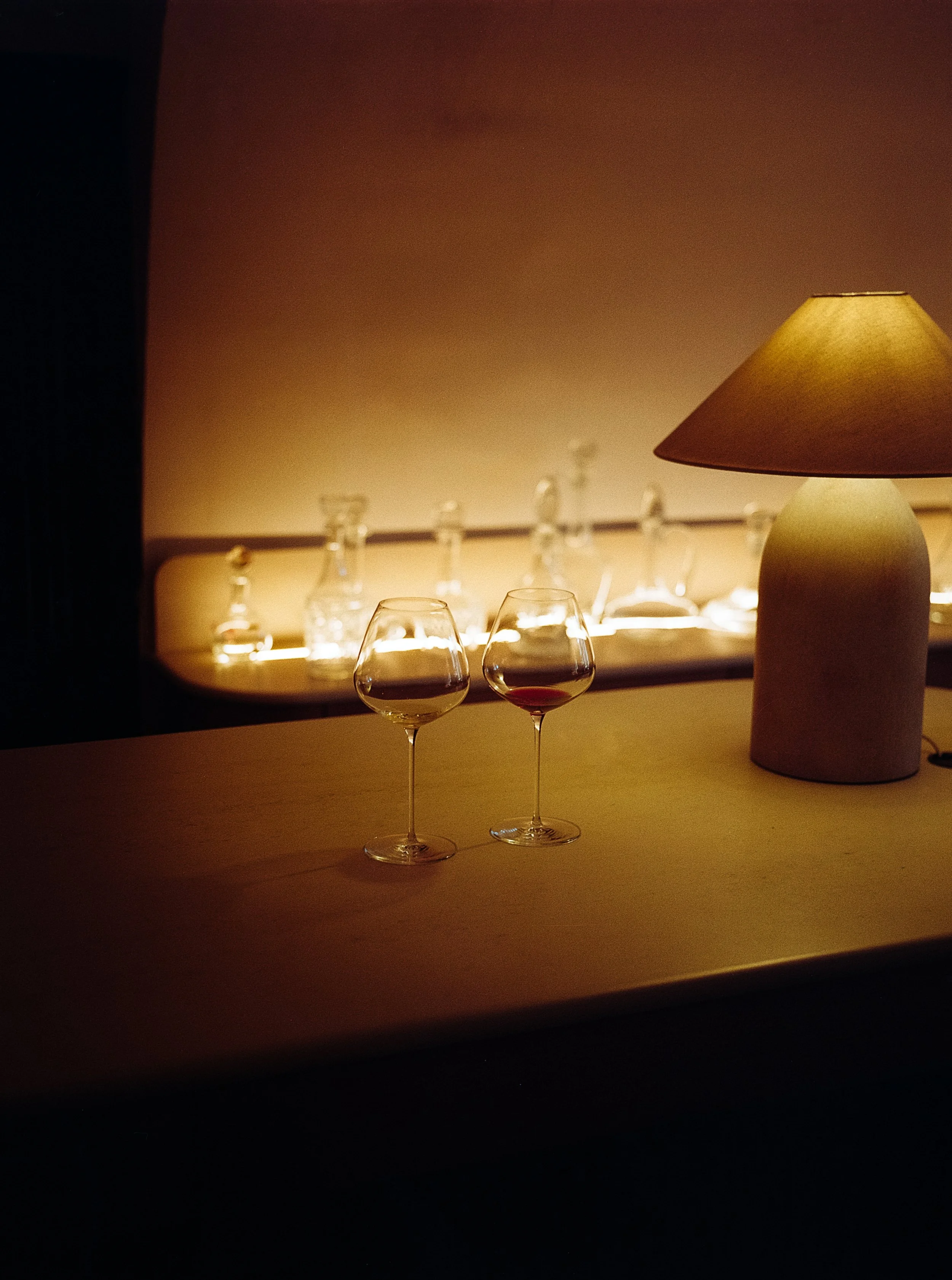 Deux verres à vin rouge sur une table en bois, éclairés par une lampe, avec un fond flou de bouteilles en verre sur une table ou un comptoir.