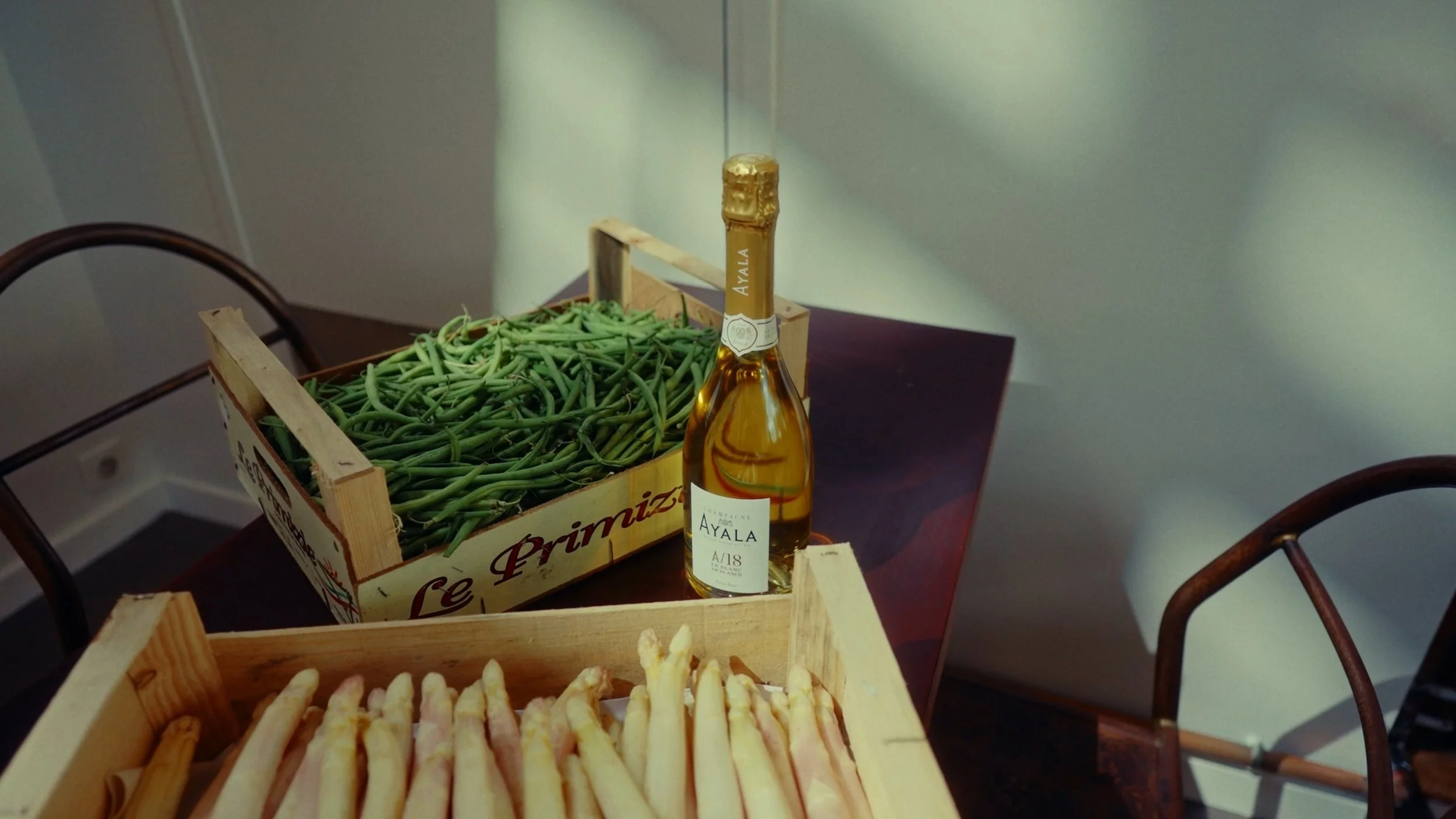Une bouteille de champagne Ayala, une caisse de haricots verts et une caisse d'asperges sur une table en bois.