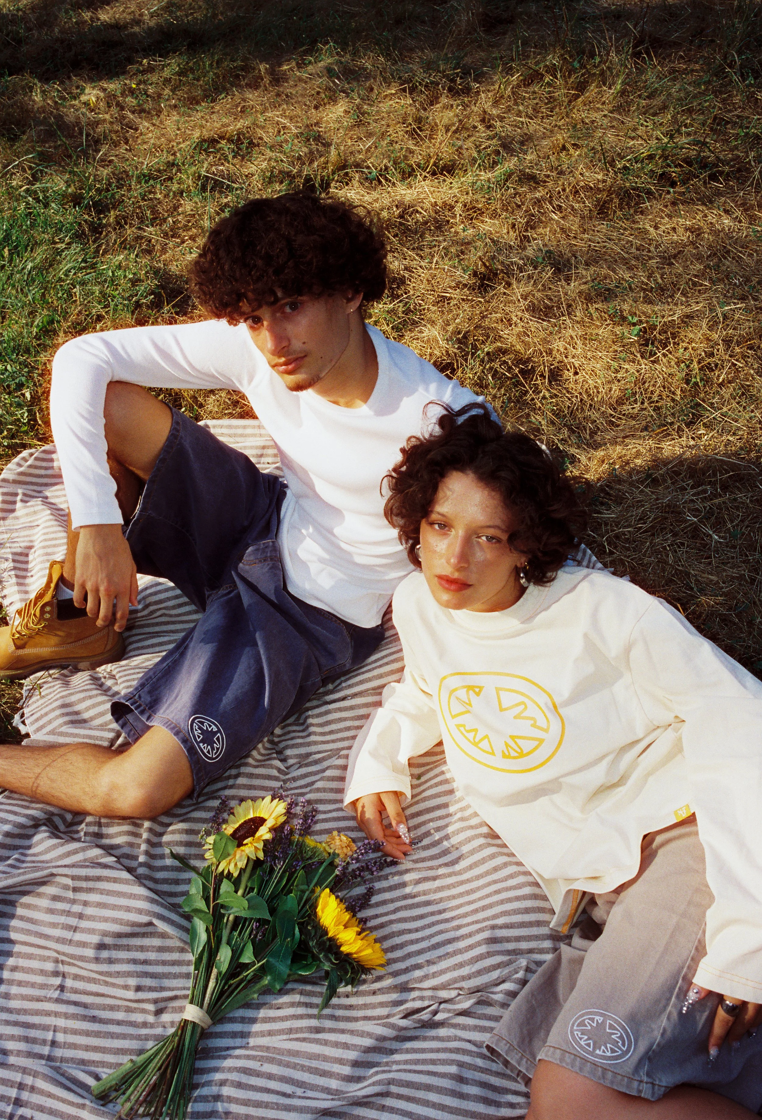 Deux jeunes adultes, un homme avec des cheveux bouclés noirs et une femme avec des cheveux bouclés bruns, allongés sur un plaid rayé en plein air avec de l'herbe jaune et verte en arrière-plan. Il y a un bouquet de tournesols posé sur le plaid.