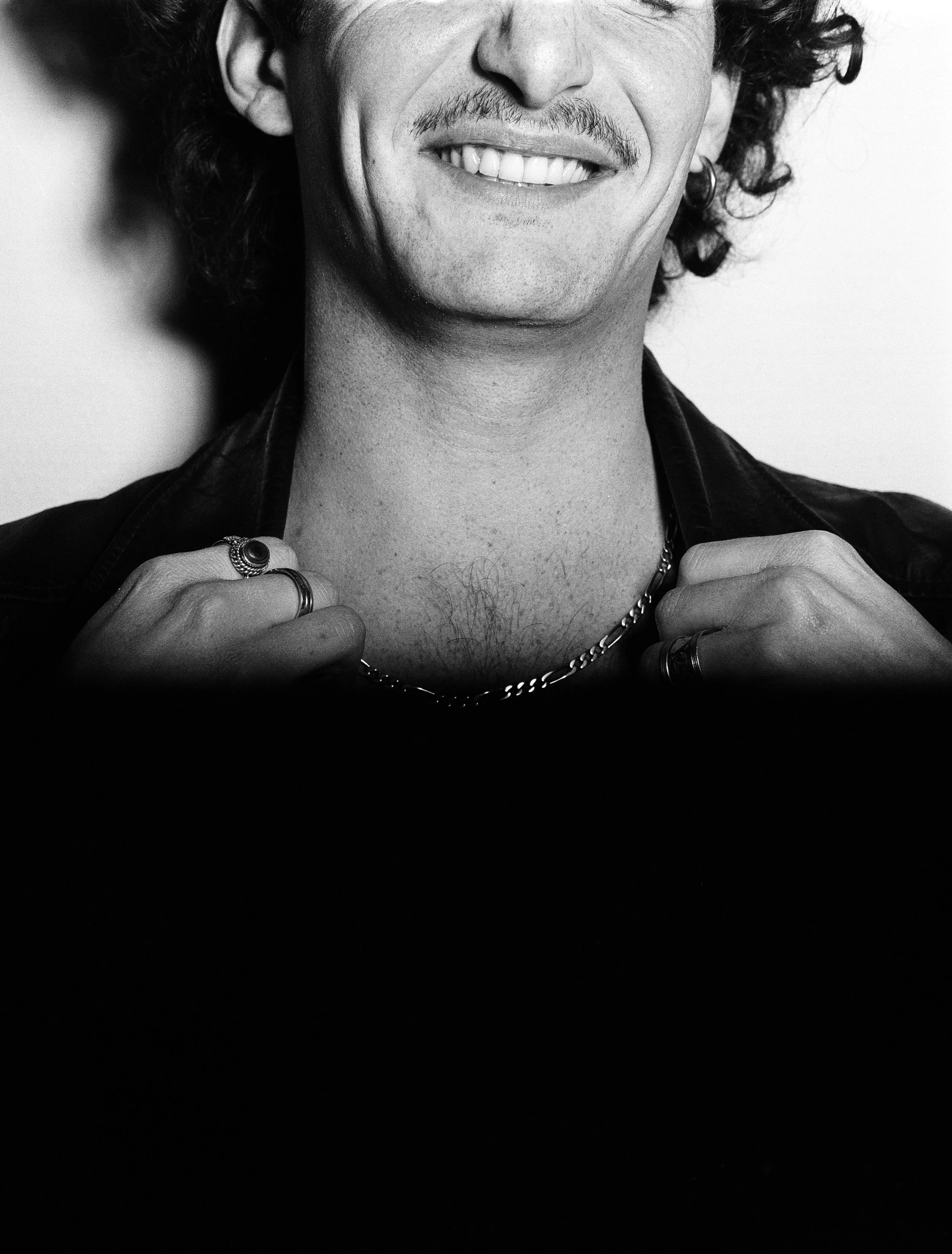 Portrait en noir et blanc d'un homme souriant, vêtu de bijoux, vêtu d'une veste en cuir.