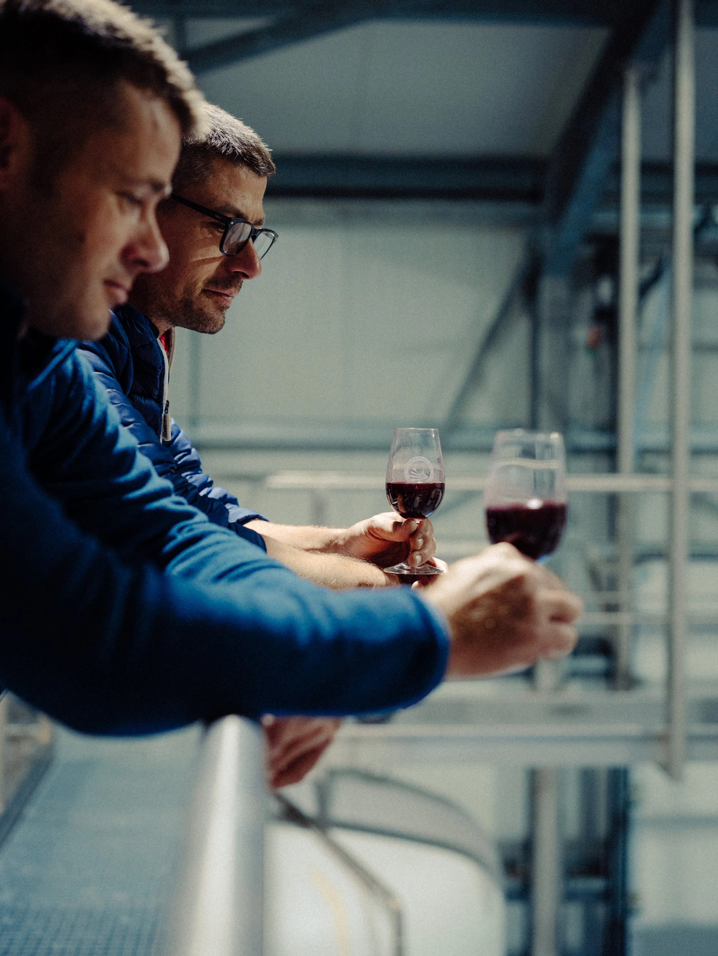 Trois hommes dégustant du vin rouge dans une cave ou une salle de production de vin.