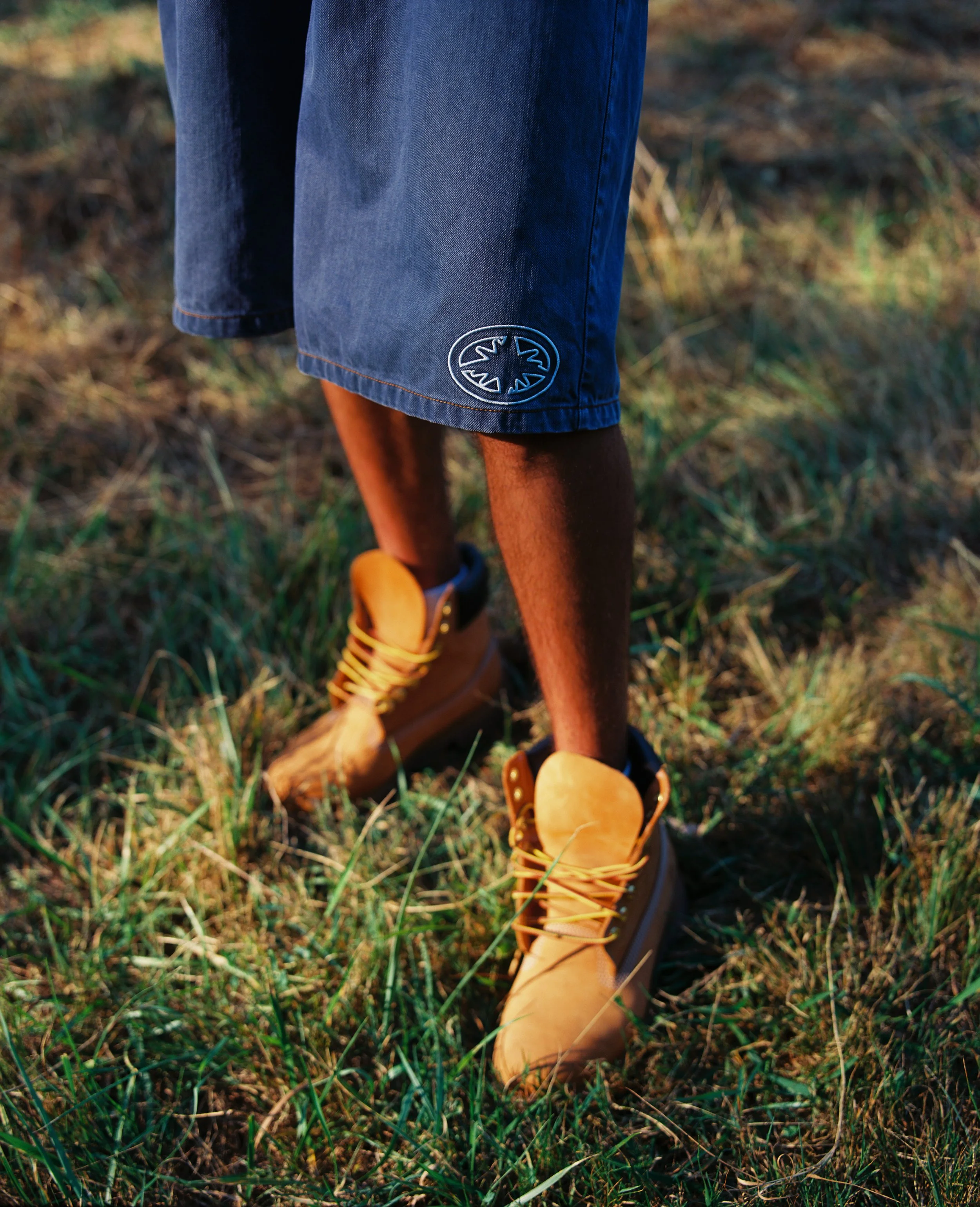 Personne portant des bottes en cuir marron et un short en denim, marchant dans l'herbe lors d'une journée ensoleillée.