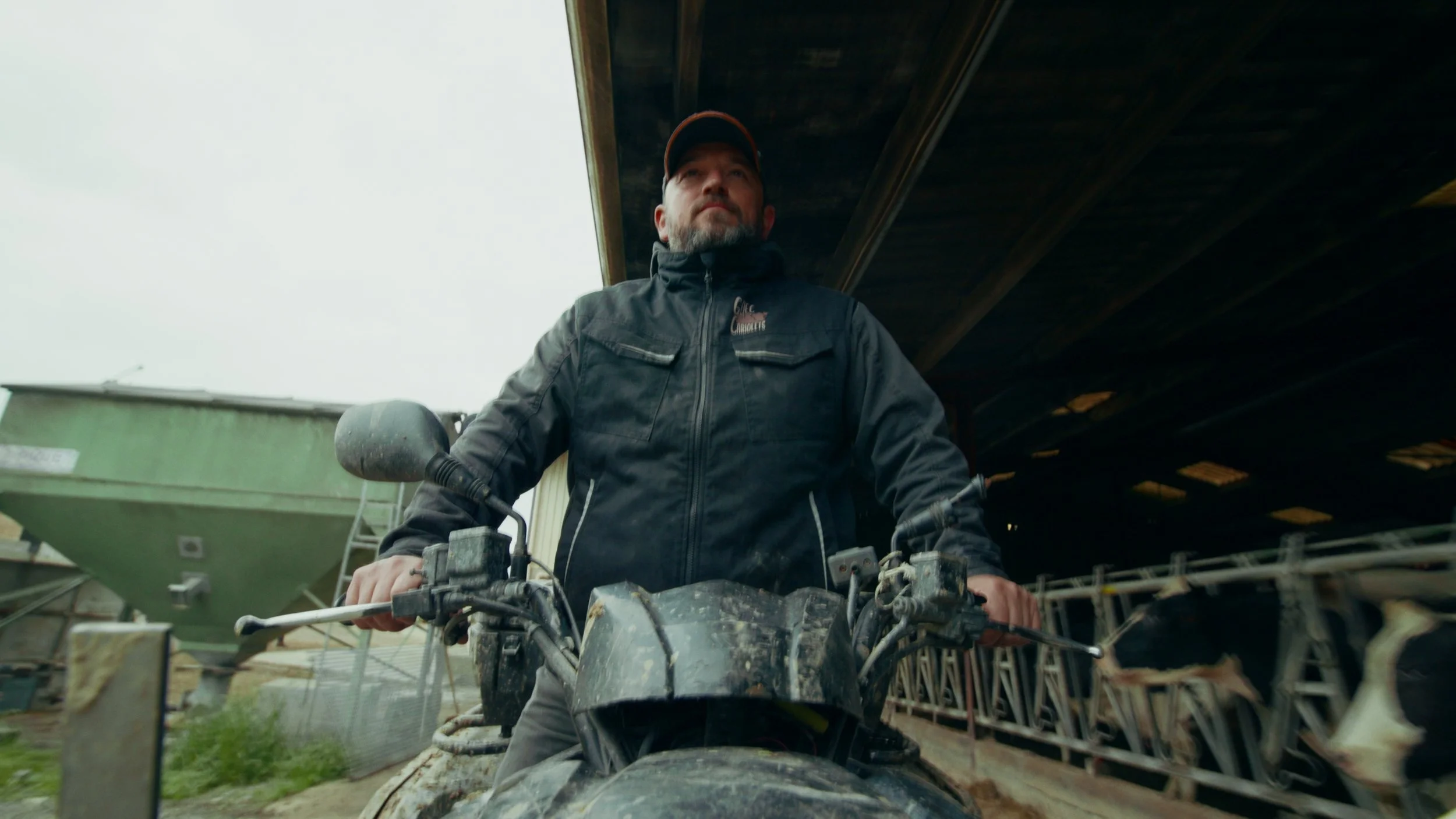 Un homme assis sur un quad, portant une veste noire, sous un grand hangar industriel.