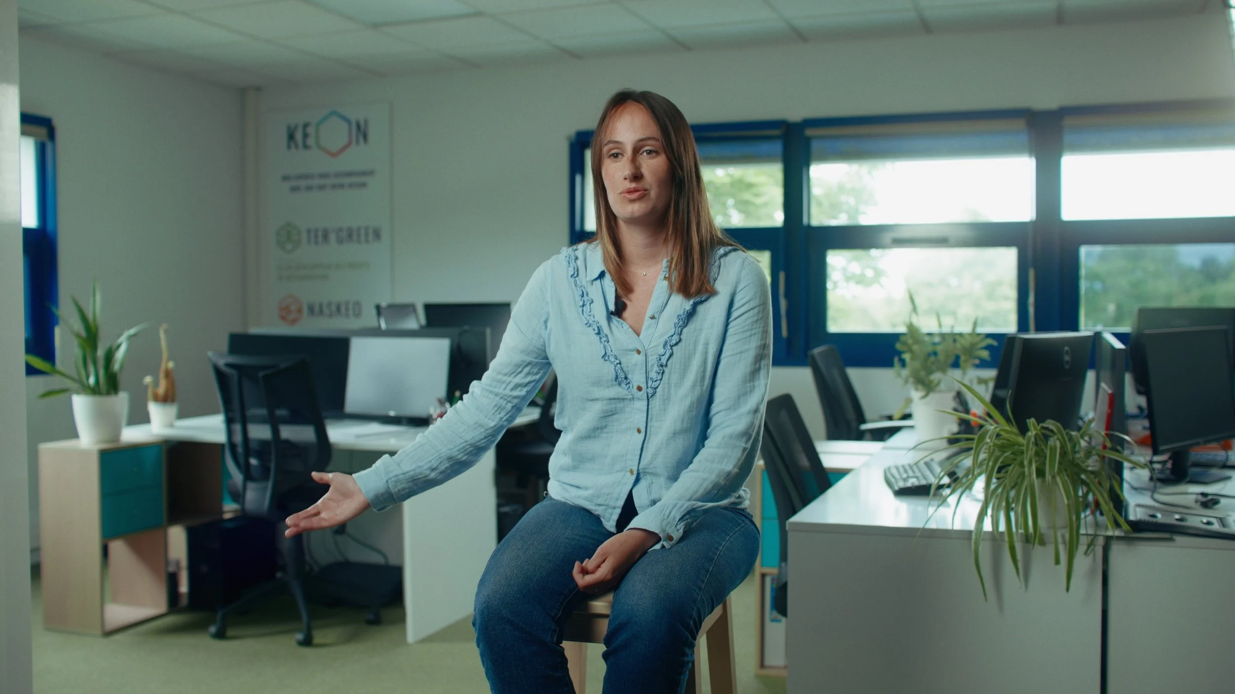 Une femme assise sur une chaise dans un bureau. Elle porte une chemise bleue et un jean, parlant ou expliquant quelque chose.