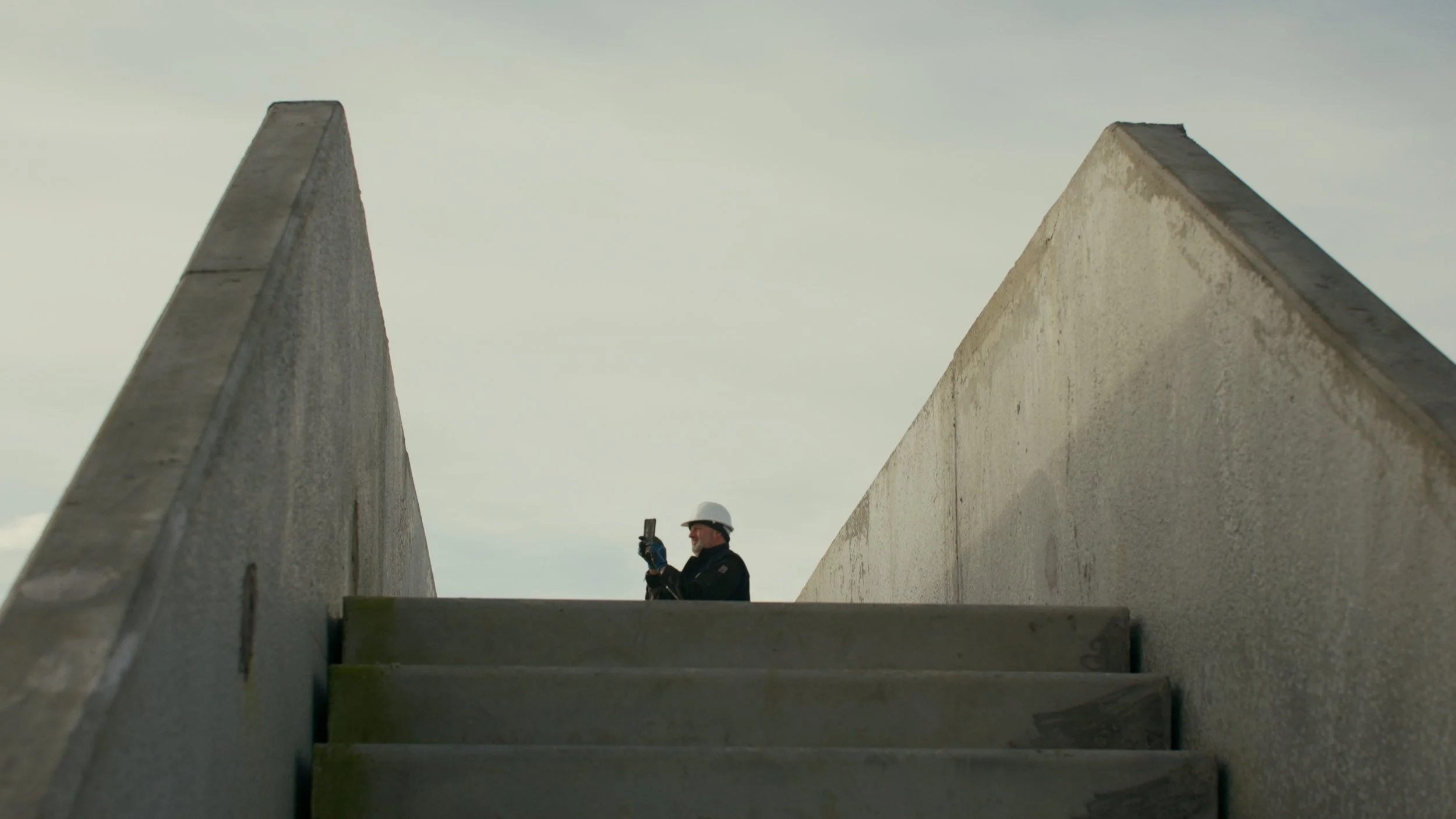 Un homme portant un casque de sécurité regarde une tablette ou un téléphone, en haut d'un escalier en béton entre deux murs.