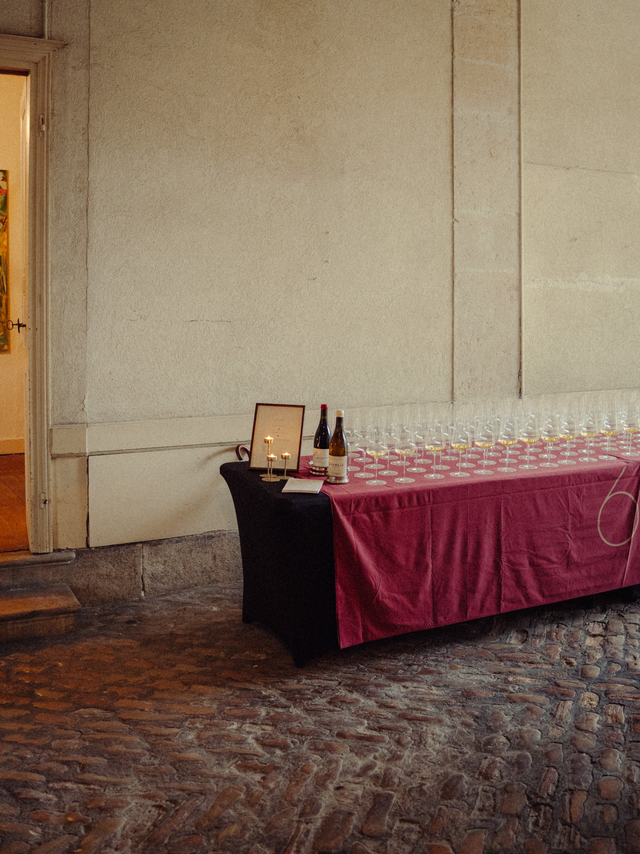 Table de dégustation avec des verres à vin, deux bouteilles de vin, un cadre, une petite bougie, et un carnet, dans une salle avec un mur beige et un sol en pavés.