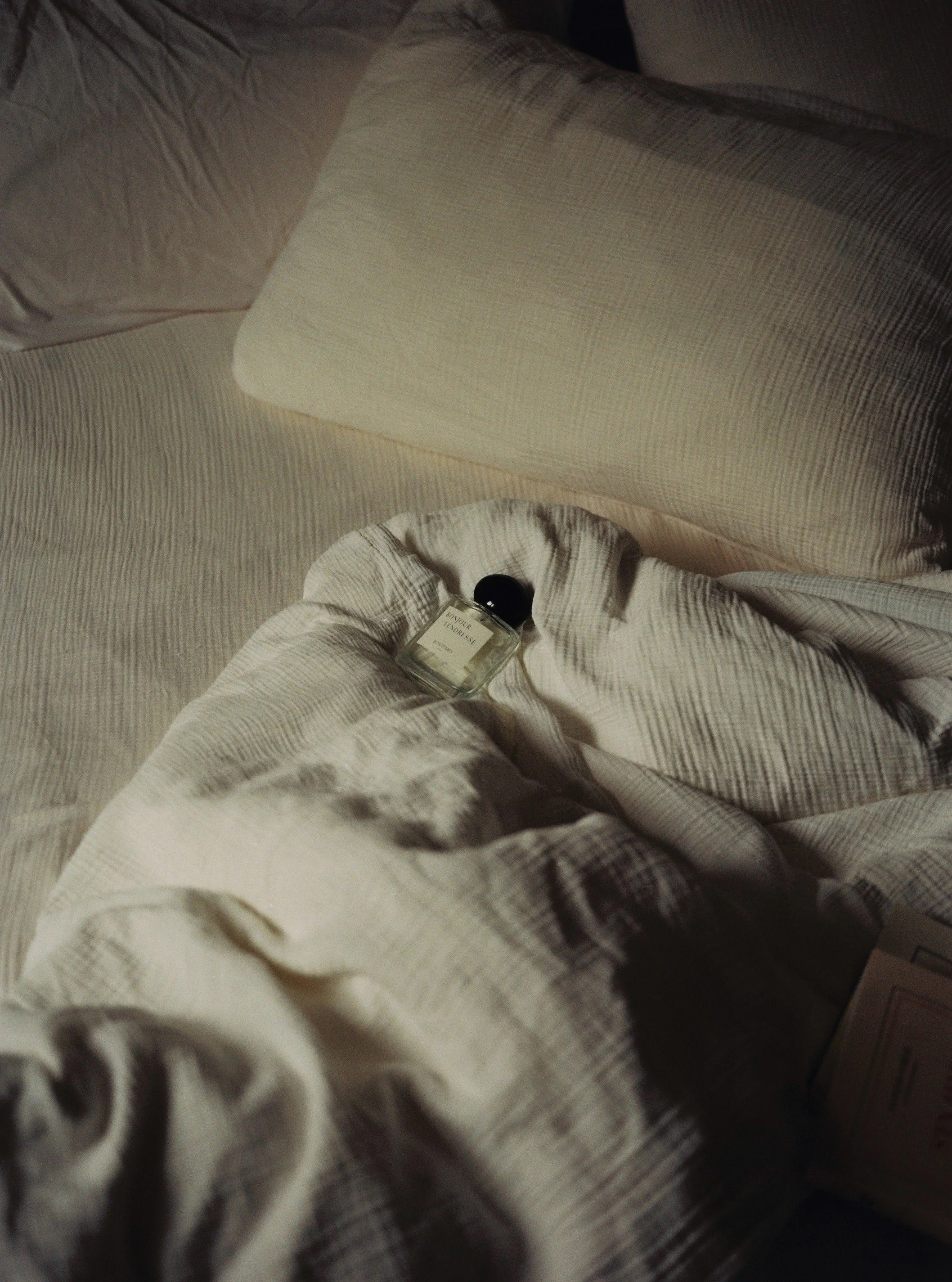 Une bouteille de parfum repose sur un drap blanc sur un lit avec plusieurs oreillers beige.