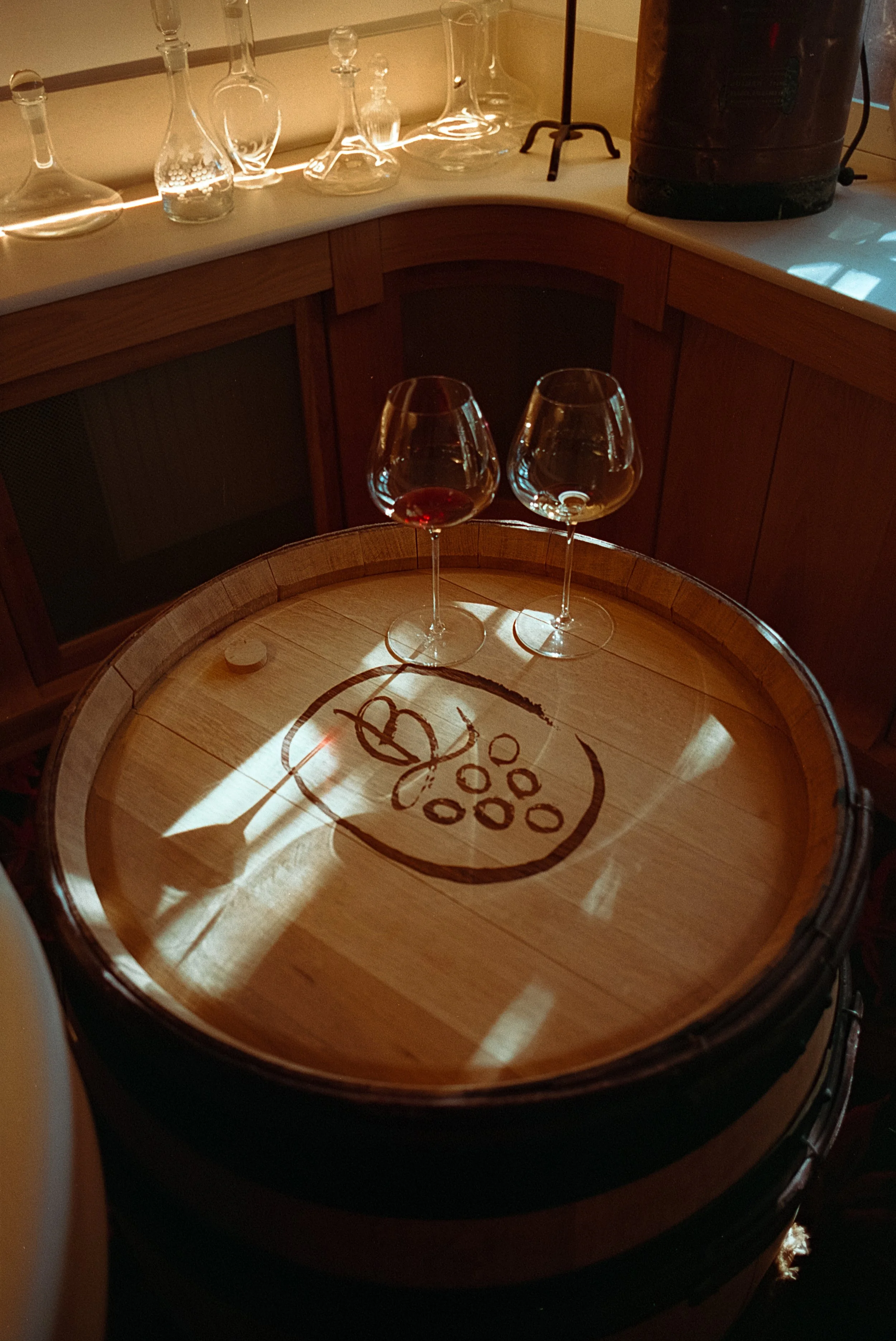 Deux verres de vin rouge posés sur une barrique en bois dans un endroit chaleureux. L'ombre de la barrique évoque un dessin d'une grappe de raisin. En fond, des bouteilles en verre et un appareil en arrière-plan.