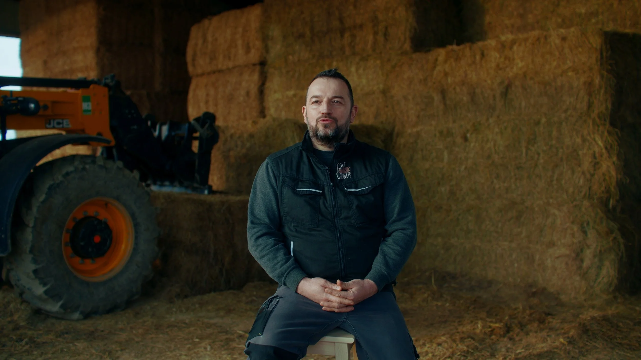 Un homme assis sur un tabouret dans une grange avec des lots de foin en arrière-plan.