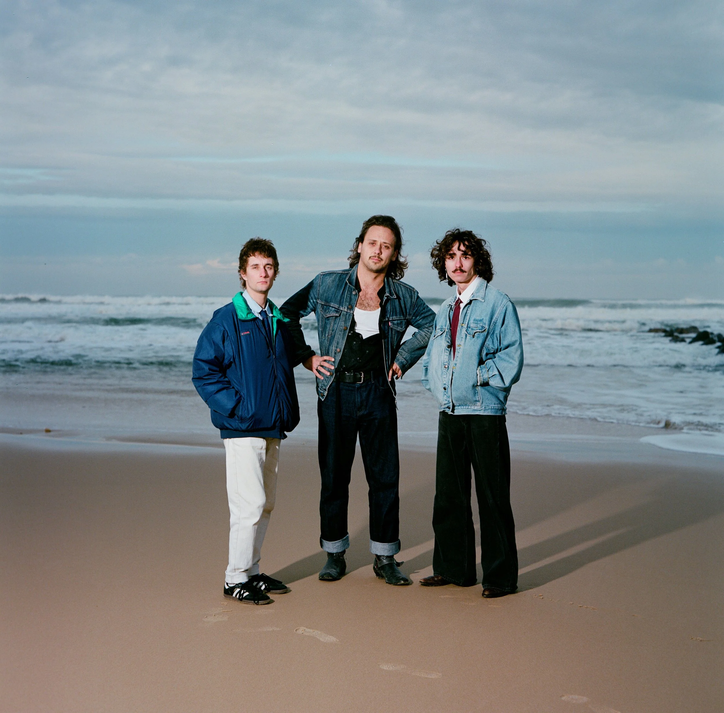 Trois hommes debout sur une plage, vêtus de vêtements décontractés, avec la mer en arrière-plan, sous un ciel nuageux.