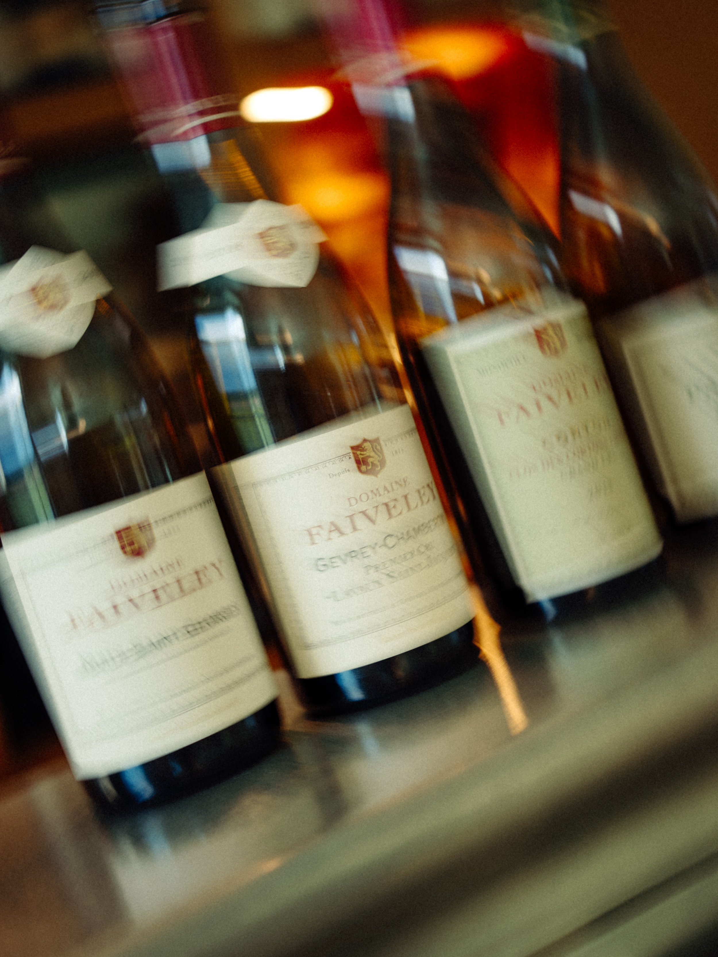 Trois bouteilles de vin en verre brun sur une étagère, étiquettes blanches avec un logo rouge et or, ambiance chaude et floue.