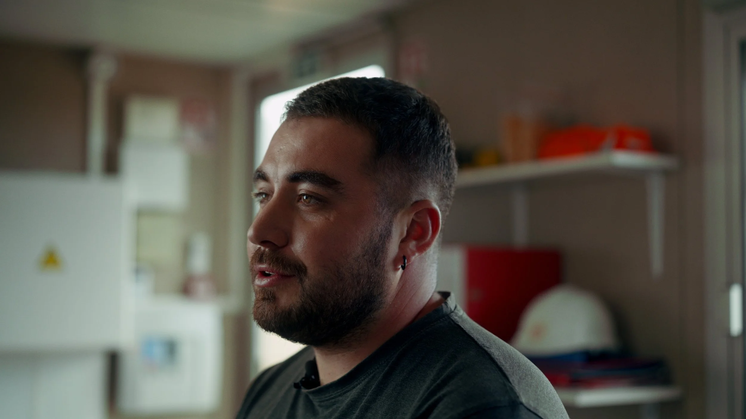 Portrait d'un homme avec une barbe, portant une boucle d'oreille, dans un intérieur avec des étagères.