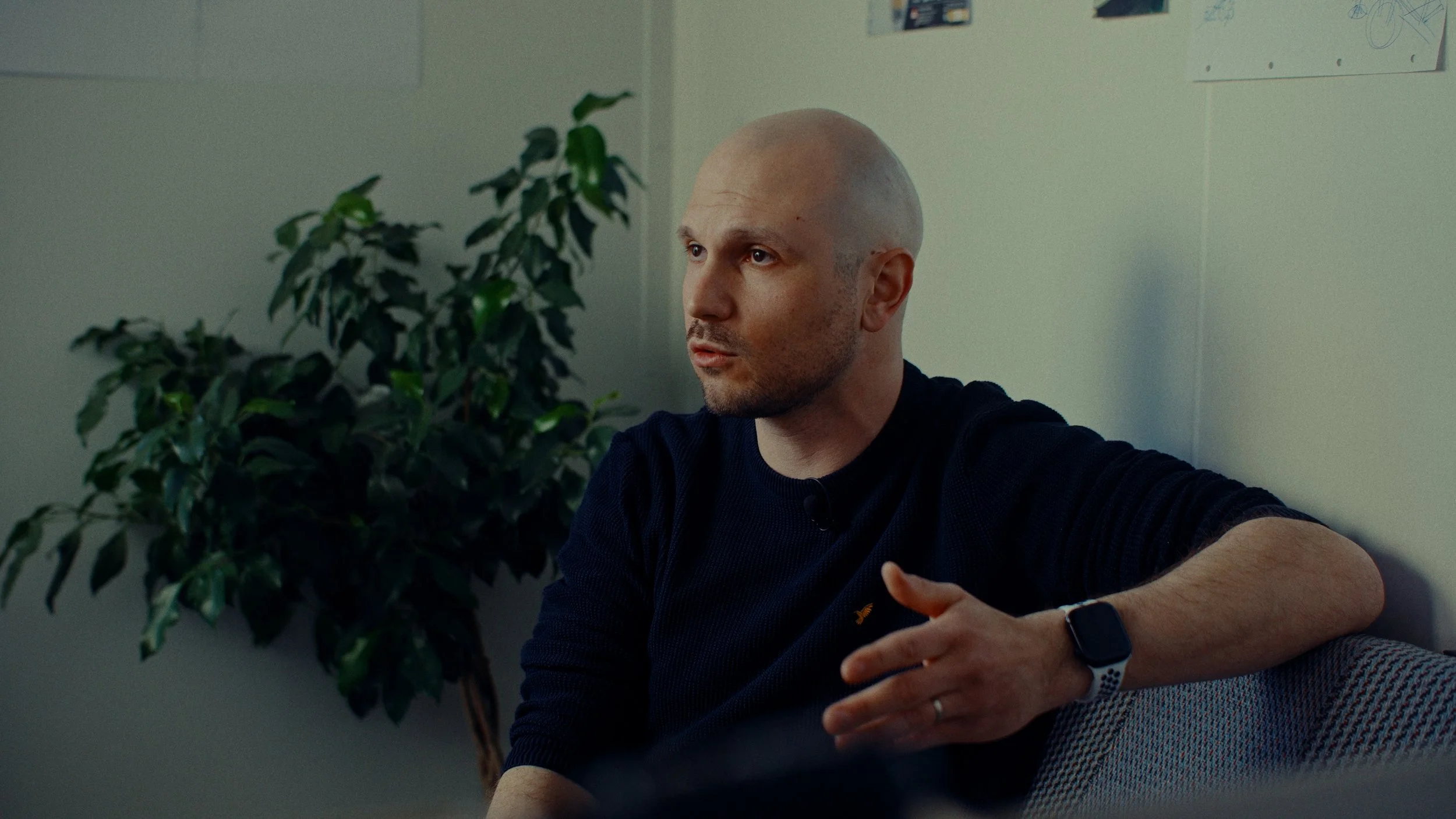 Homme parlant lors d'une interview en intérieur, portant une montre connectée, assis sur un canapé avec une plante verte en arrière-plan.