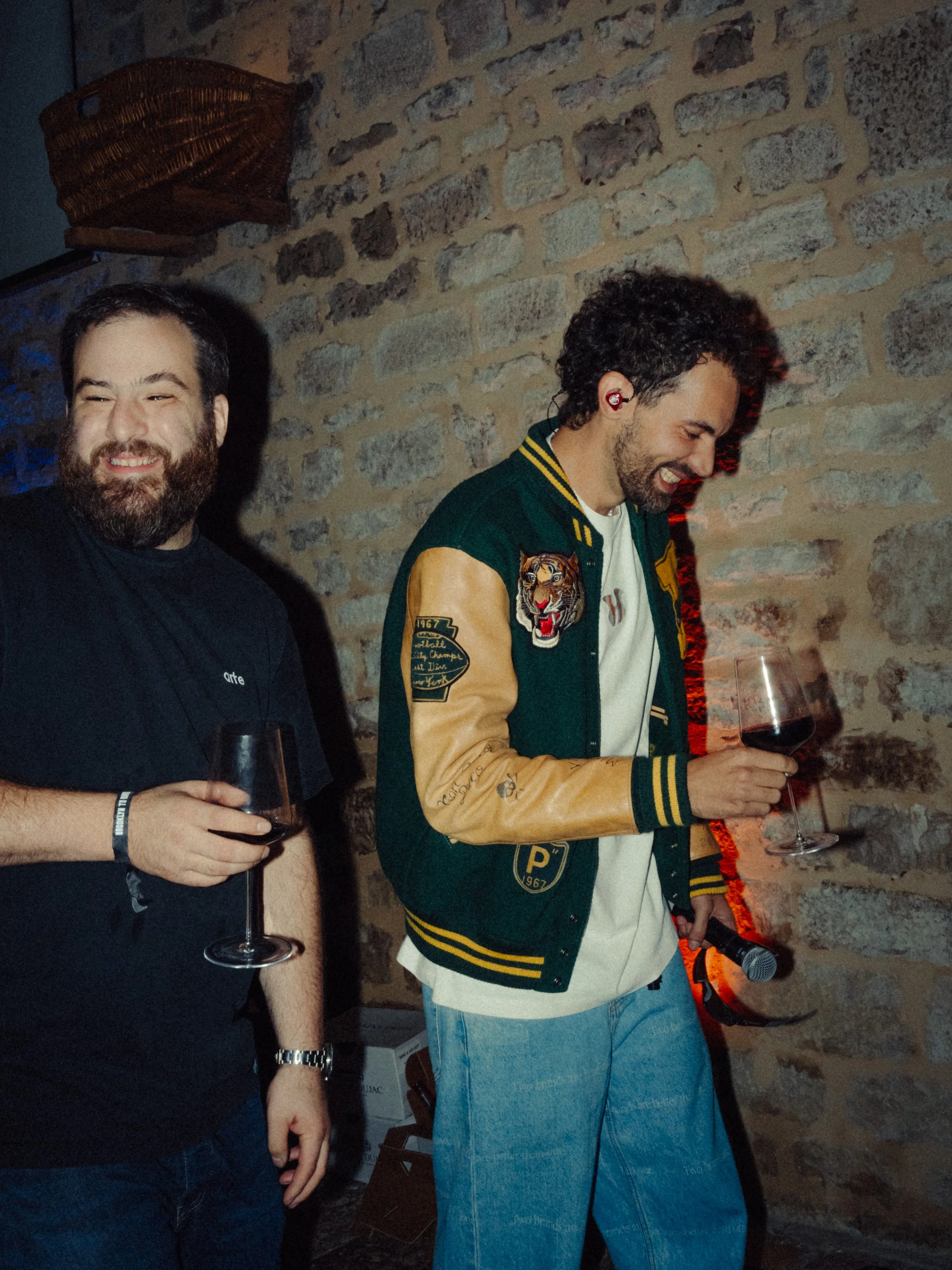 Deux hommes souriants, tenant des verres de vin rouge, dans une ambiance décontractée avec un mur en pierre en arrière-plan.