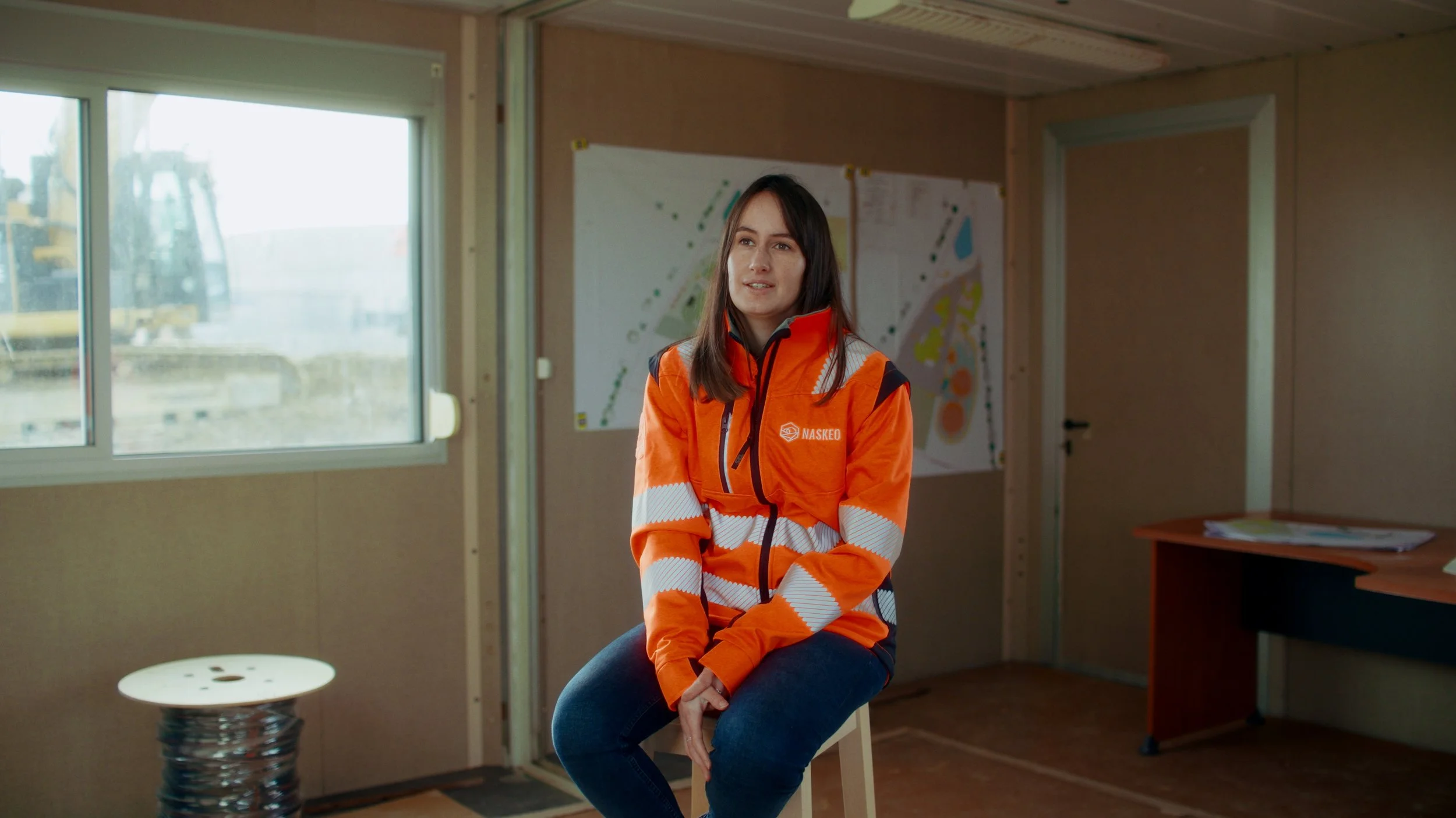 Une femme portant une veste orange avec des bandes blanches, assise sur un tabouret dans une pièce avec des plans et un bureau.