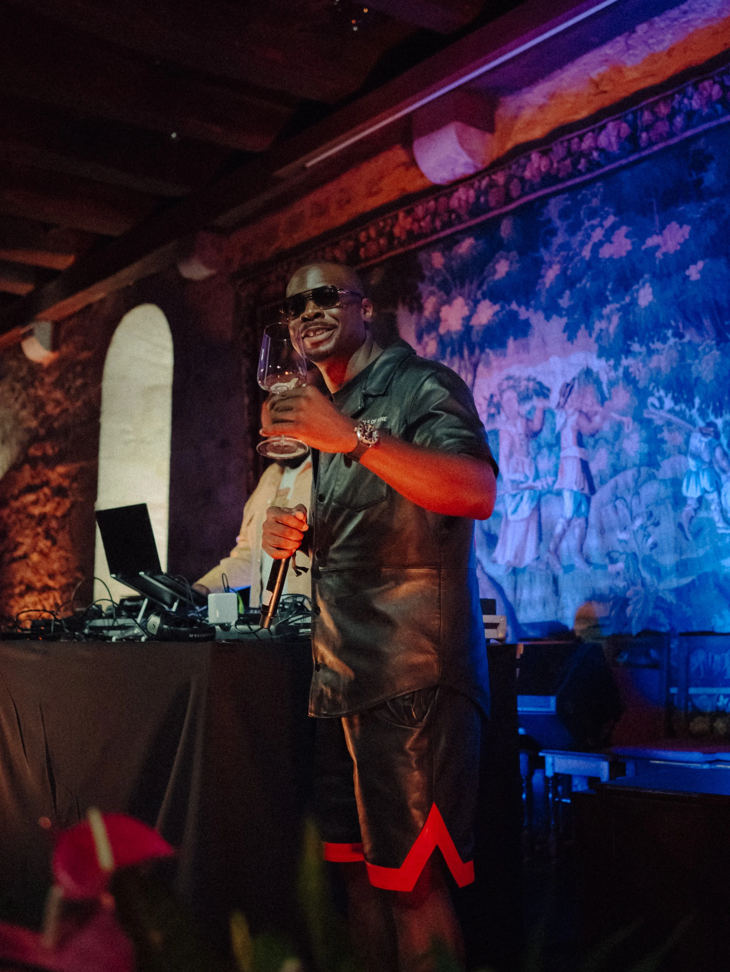 Un homme souriant portant des lunettes de soleil, un cuir noir, tenant un verre à vin rouge dans une main et un microphone dans l'autre, lors d'une performance ou d'une fête dans une ambiance tamisée avec une décoration artistique en arrière-plan.