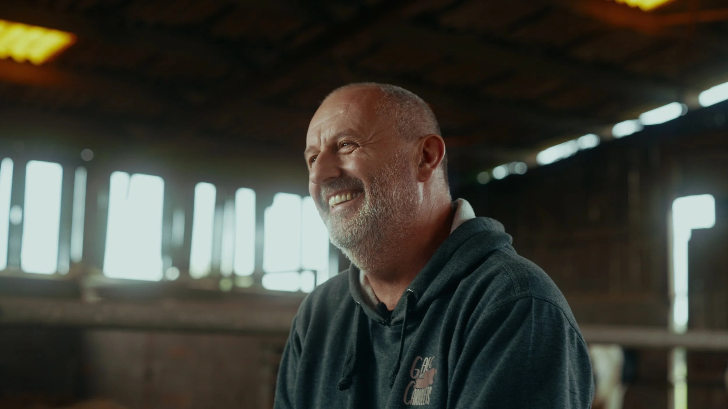 Un homme âgé avec une barbe et un sourire, portant un sweat à capuche gris, dans une ferme ou une grange en bois, avec des fenêtres laissant passer la lumière naturelle.