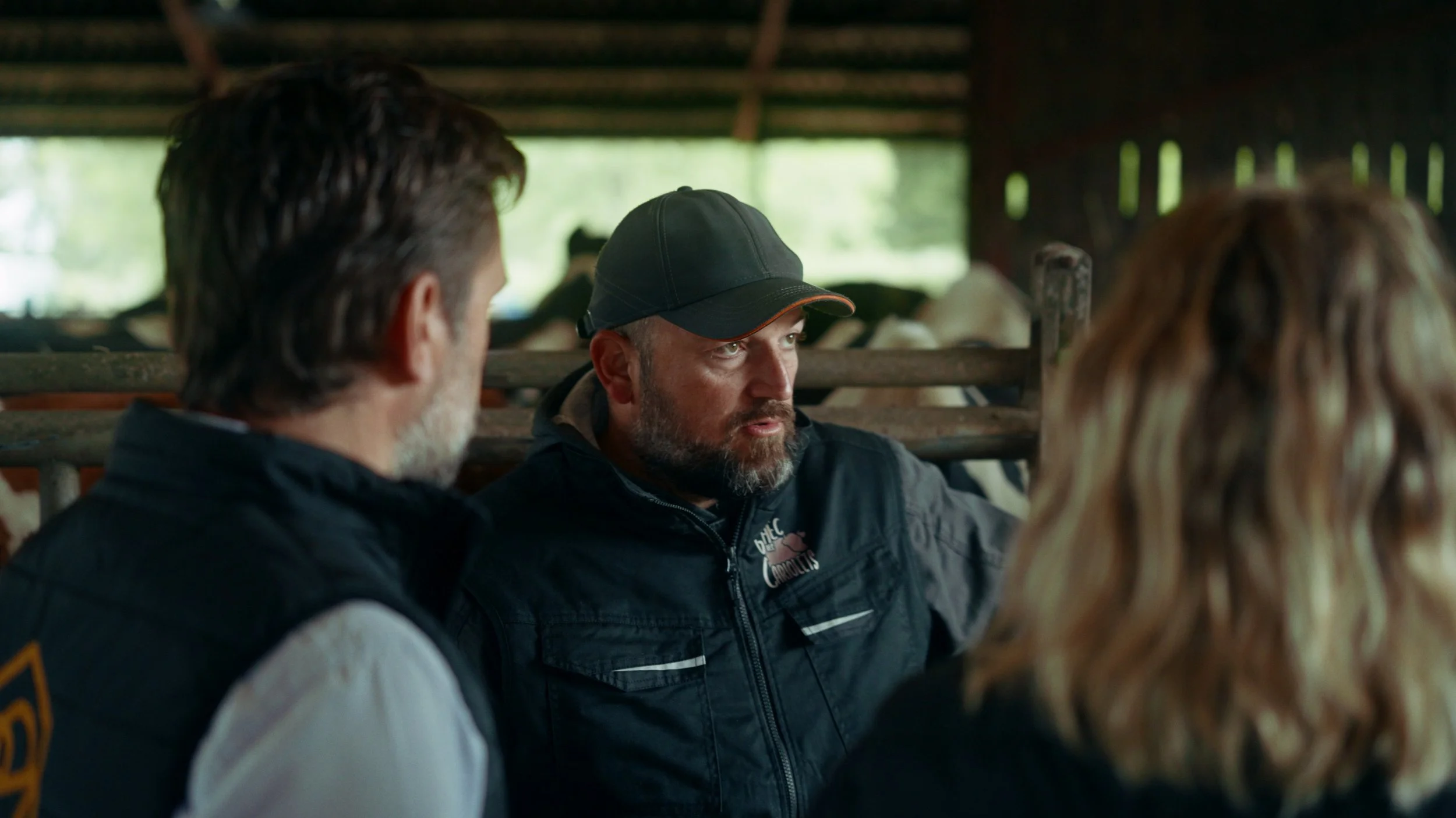 Trois personnes discutent dans une ferme avec des vaches en arrière-plan.