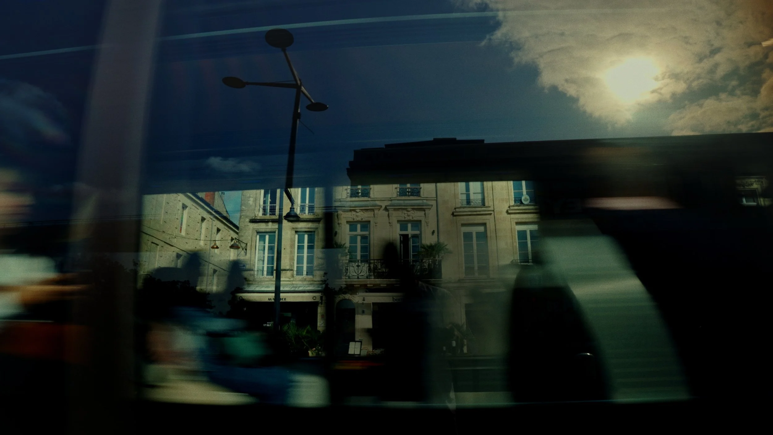 Une vue urbaine avec des bâtiments historiques, une rue, un lampadaire moderne, et un ciel ensoleillé avec quelques nuages, vue à travers une surface réfléchissante ou une fenêtre.