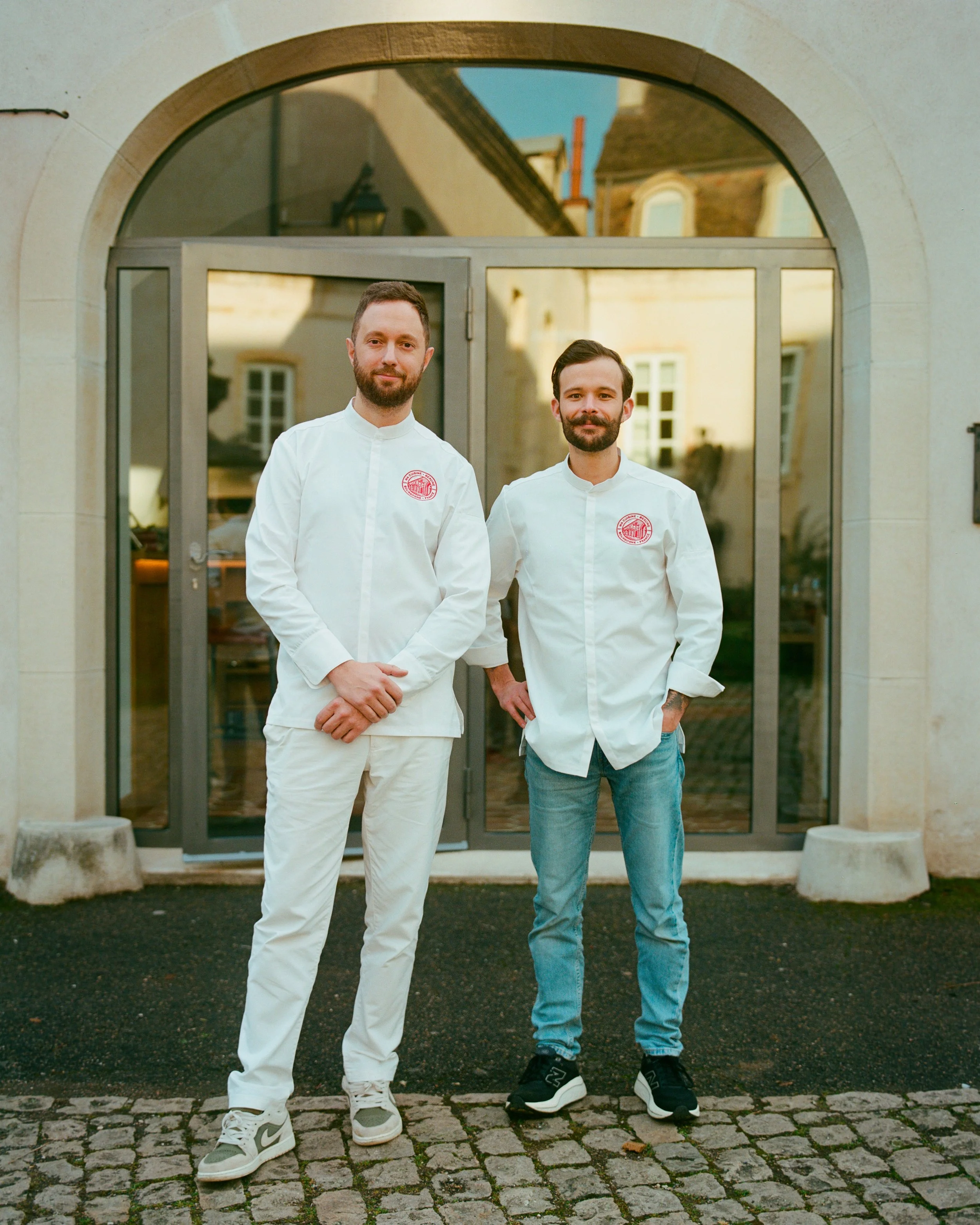 Deux hommes debout devant une entrée de bâtiment, portant des uniformes de cuisine blancs avec un logo rouge, souriants, l'un a les bras croisés, l'autre la main dans la poche, en extérieur sur un sol pavé.