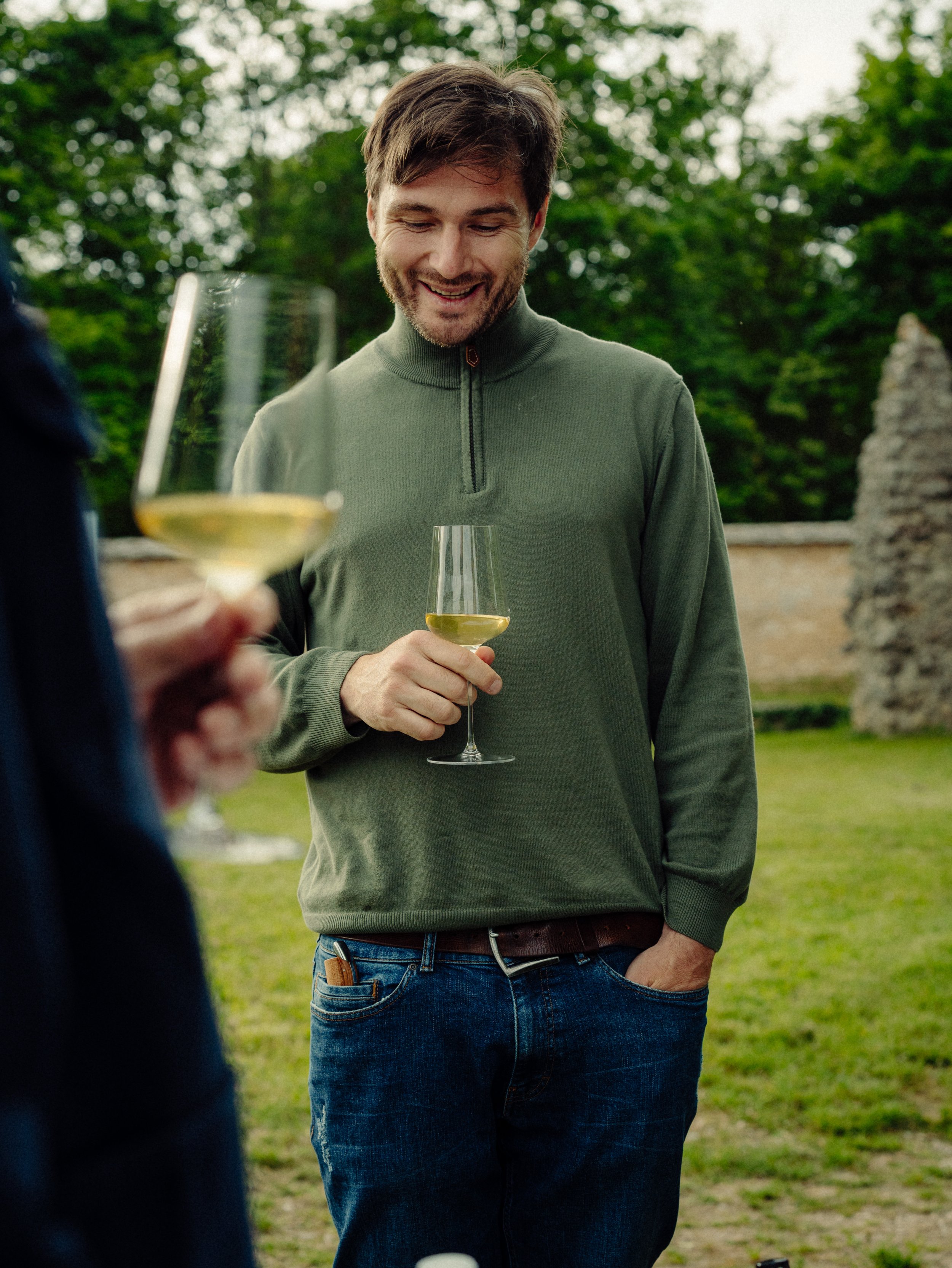 Un homme souriant qui tient un verre de vin blanc dans une main, lors d'une dégustation en plein air, avec un décor de verdure et un mur en pierre en arrière-plan.