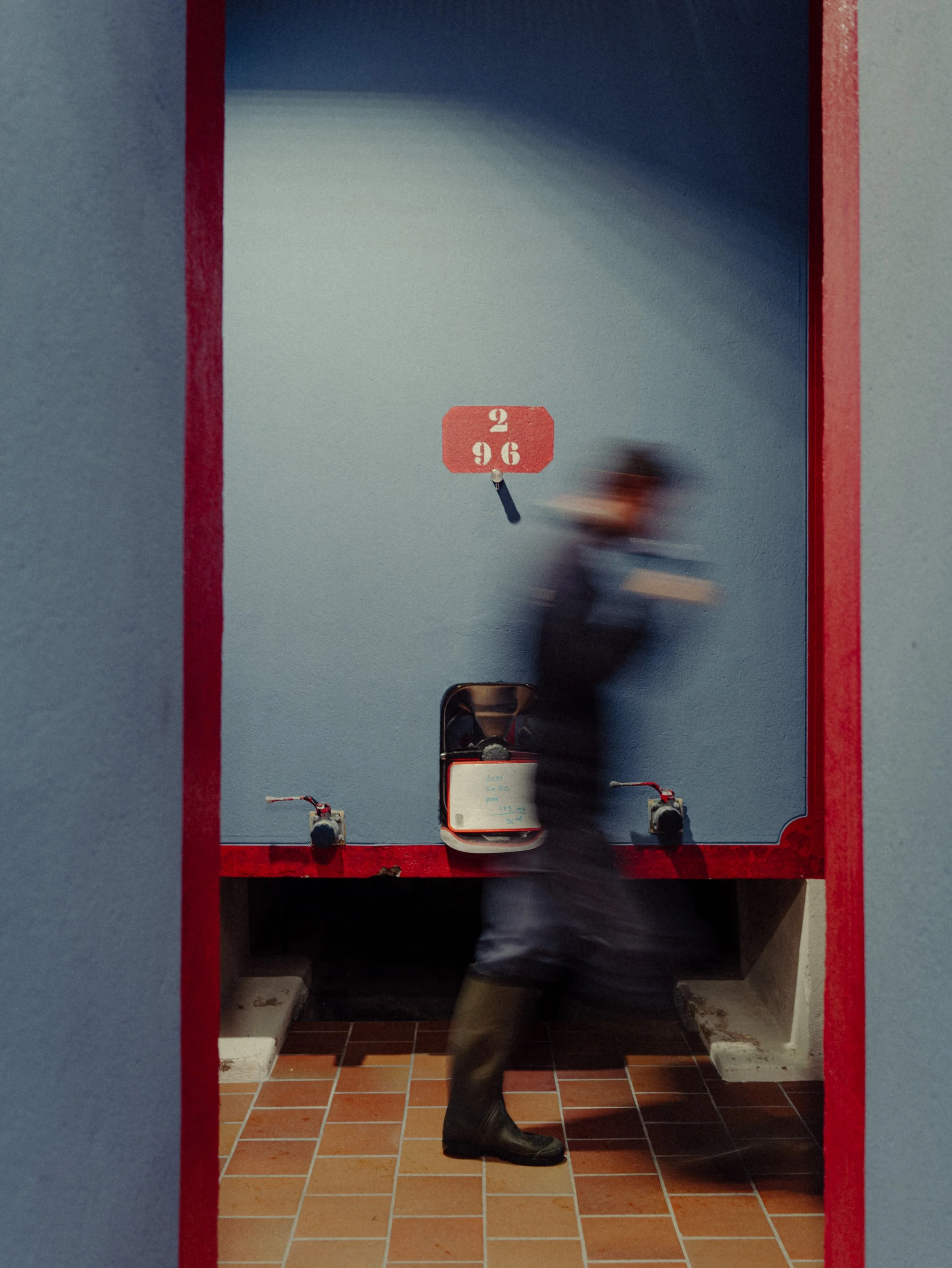 Une personne en déplacement, debout dans une zone légèrement sombre, dans une pièce avec un grand tableau ou panneau blanc, affichant une horloge ou un compteur dont les aiguilles pointent vers 2h et 96, avec deux petits appareils ou éléments en bas 