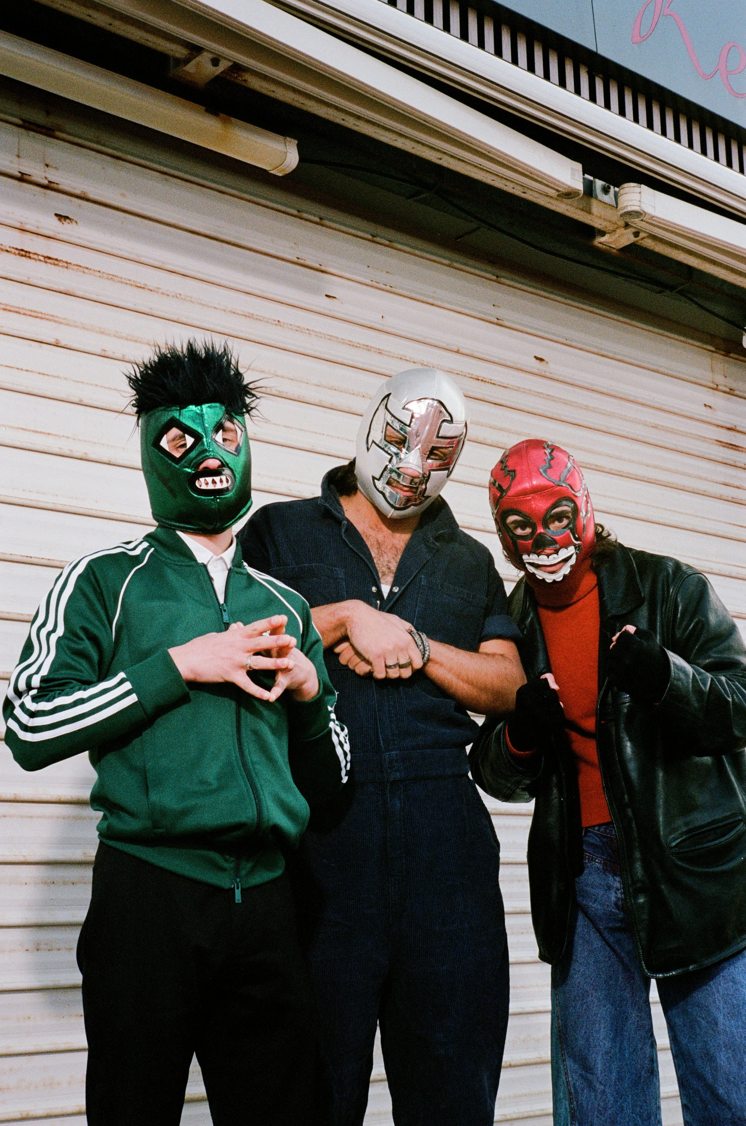 Trois personnes portant des masques de lucha libre mexicaine côte à côte devant une porte en métal.
