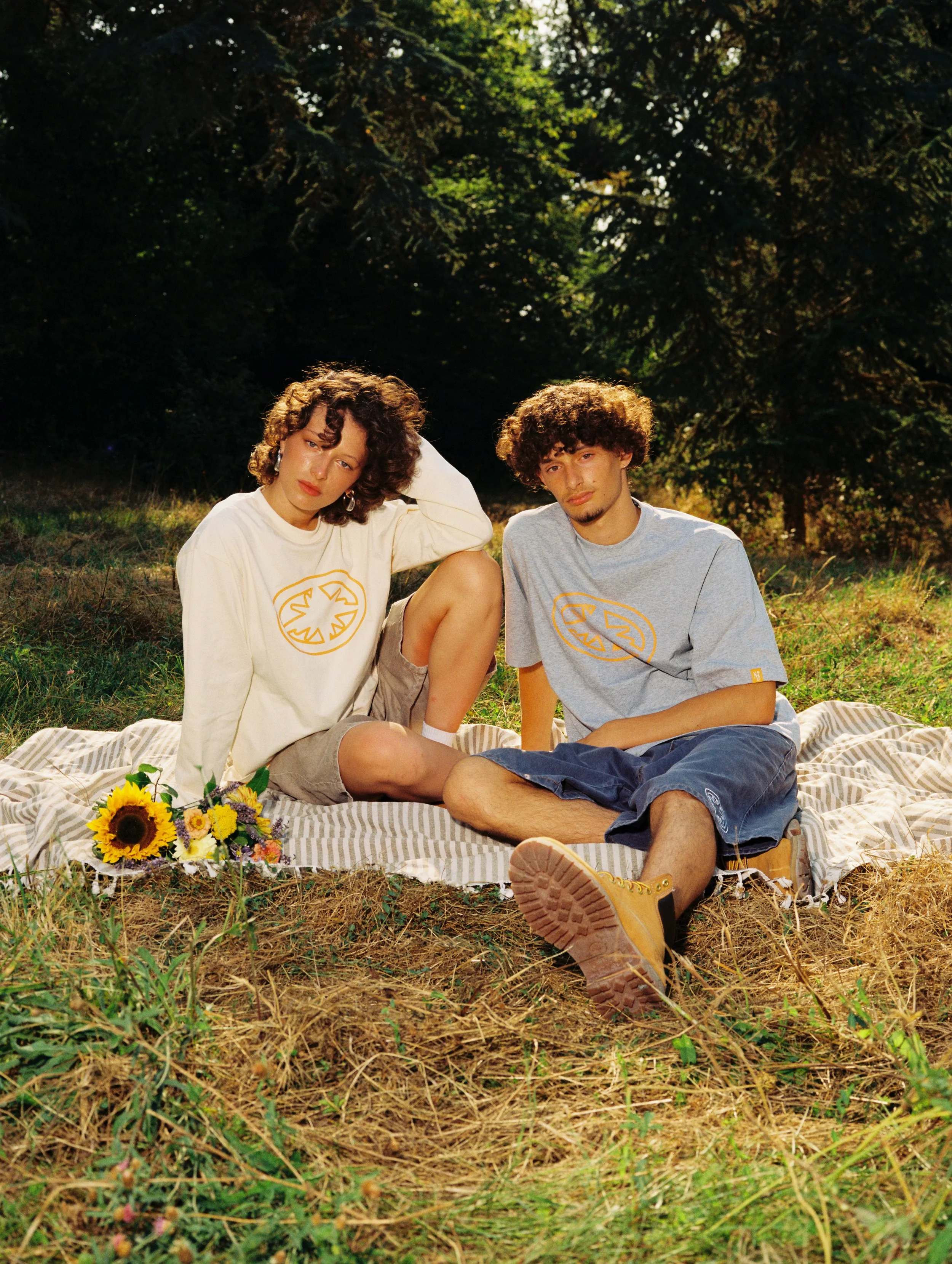 Deux jeunes hommes et une jeune femme assis sur une couverture dans une forêt, entourés de verdure et de soleil, avec un bouquet de tournesols posé à côté.